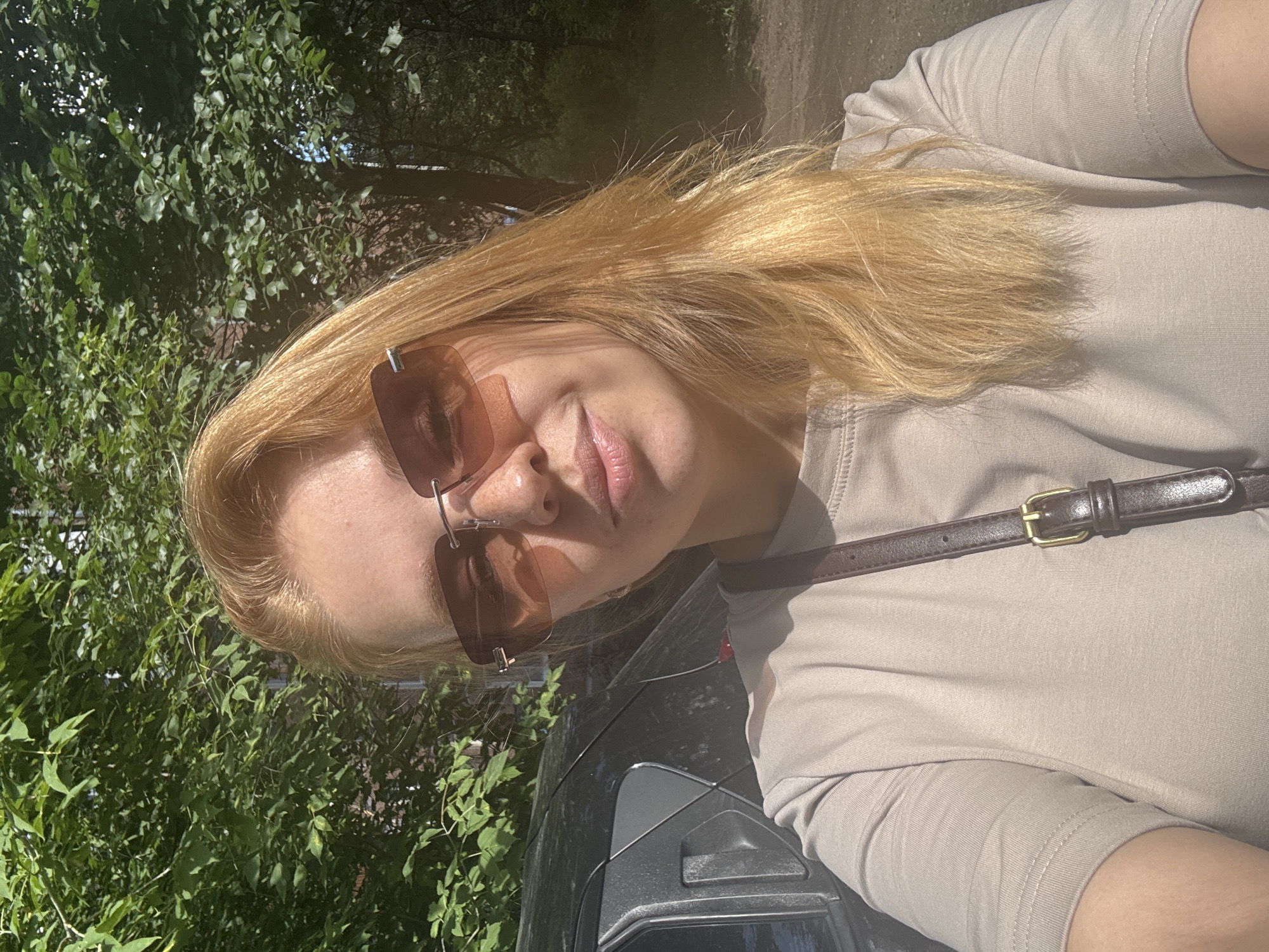 A woman with blonde hair and sunglasses is taking a selfie outdoors. She is wearing a light-colored t-shirt and a dark brown strap is visible across her chest. Behind her, a dark-colored car is parked next to a brick building, partially obscured by green trees. The scene suggests a sunny day.