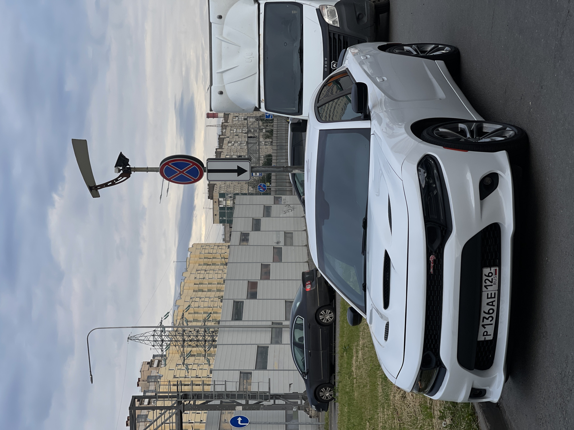 The image captures a street scene in an urban environment under an overcast sky. In the foreground, a white Dodge Charger is parked, facing slightly to the right. The car features a prominent hood scoop and a license plate reading "P136AE 126" with "RUS" below it. To the left of the Charger, a dark-colored sedan is visible. Behind these cars, a white commercial van is parked, its front end partially obscured.
A busy road separates the vehicles from a large, light-colored building complex in the background. This complex appears to be a residential or commercial area with multiple stories and many windows. Industrial elements, such as tall power lines and transmission towers, are also present in the distance, suggesting an area that combines urban living with infrastructure.
A traffic signal pole stands to the left of the van, displaying a circular "no stopping" sign with a red border and a blue background with a red X. Below this, a rectangular sign with a black arrow pointing downwards is mounted. A blue and white directional sign indicating a right turn is visible further left. The environment is paved with asphalt, and a strip of green grass runs along the side of the road where the Charger is parked. The overall impression is of a typical city street, possibly during the daytime given the lighting, though the cloudy sky suggests subdued light conditions.