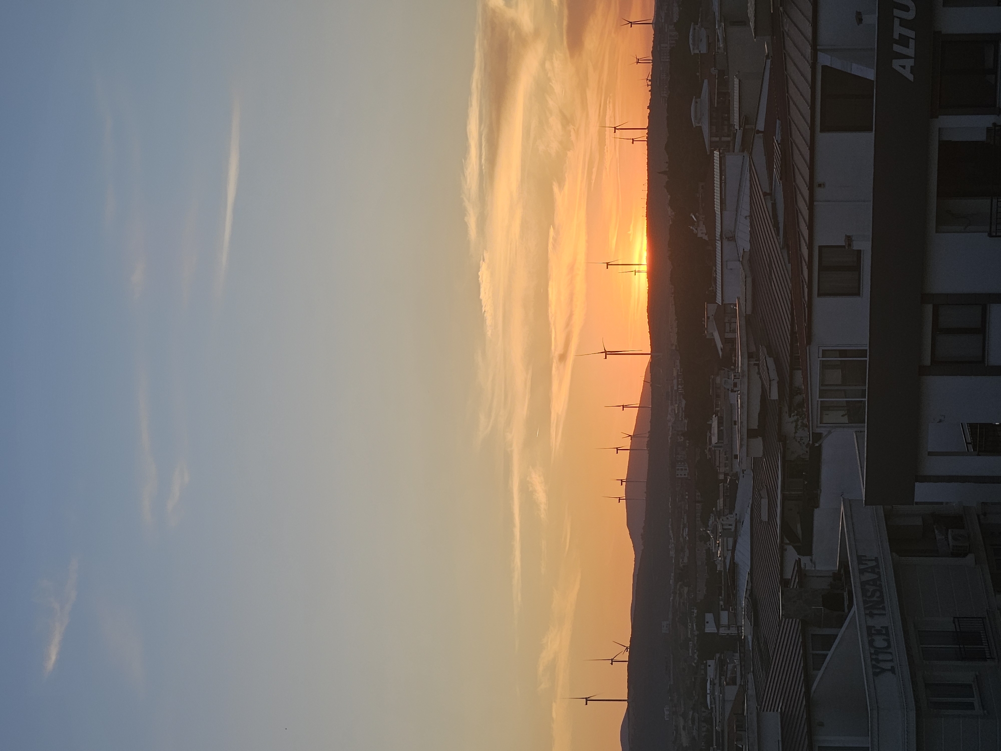 The image depicts a panoramic view of Seferihisar, Turkey, at sunset. The sky is a gradient of soft blues transitioning into vibrant oranges and yellows, illuminated by the setting sun. Scattered clouds, some wispy and others more defined, are visible in the sky.
Dominating the middle ground are numerous wind turbines positioned on a silhouetted hill range. These turbines are a prominent feature of the landscape, suggesting a renewable energy presence in the region.
In the foreground, the rooftops of residential and commercial buildings are visible. The architecture appears to be of modern construction, with various roof materials and building styles. Some buildings have visible signage; on one building, the text "YÜCE İNSAAT" is clearly legible, while on another, the partially visible word "ALTU" suggests a commercial establishment.
The overall scene conveys a sense of a coastal city or town transitioning from day to night, with the natural beauty of the sunset complemented by the man-made structures and the industrial presence of the wind farm. There are no people or animals visible in the frame. The environment is characterized by the urban landscape set against a dramatic natural sky.