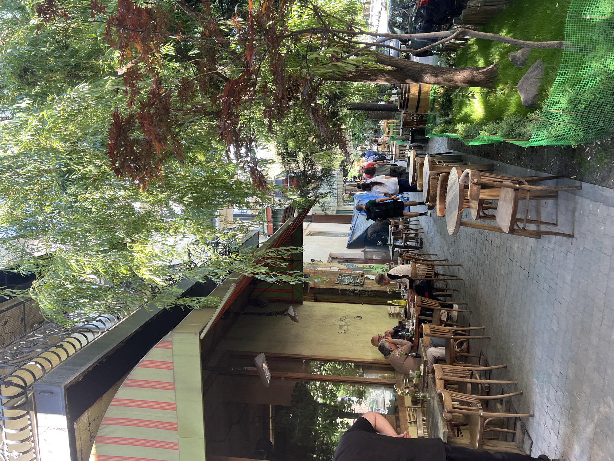 The image captures a street-level view of an outdoor dining area, presumably in Yerevan, Armenia. The scene is shaded by trees, some with reddish-brown leaves. Along the right edge of the frame, tables and chairs are arranged on a brick-paved surface. Patrons are seated at some of the tables. Some people are standing among the tables, one is in black clothing. Adjacent to the tables, there is a small patch of greenery. A car is parked beyond the green patch. On the left, the awning of a building with striped fabric is visible. The awning is attached to a building. People can be seen seated inside. The writing "SURP GHUKAS" is visible on the building's wall.