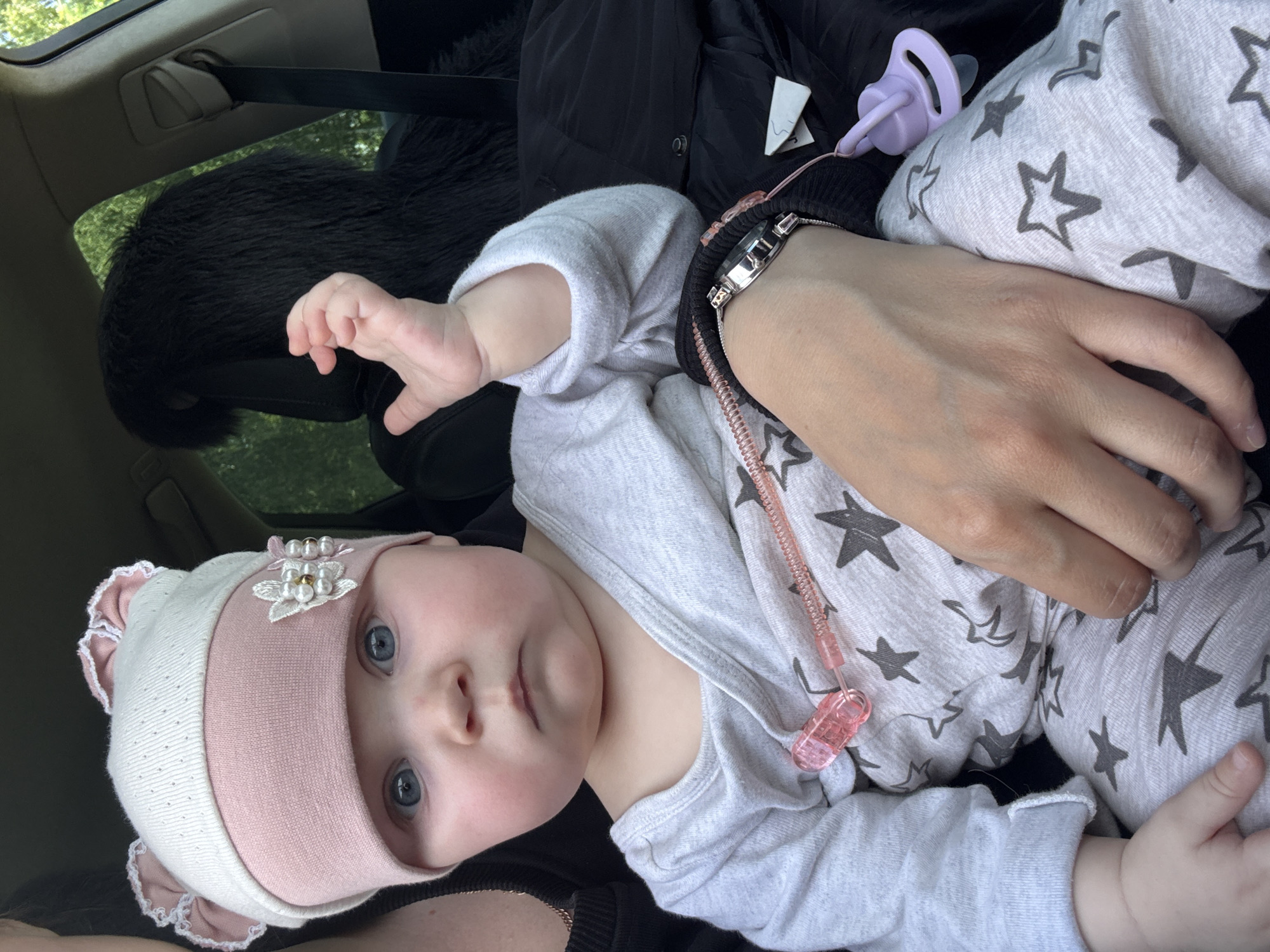 The image shows a baby with blue eyes lying in what appears to be a car. The baby is wearing a white and pink hat with a floral decoration and a light gray and white star-patterned onesie. An adult's arm and hand are visible holding the baby. A woman's watch is on the wrist of the adult. A pacifier is visible near the baby. The background includes a dark car seat and a glimpse of trees outside the window.
The location is not discernible. No part of the image provides any clue to the location within the city of Votkinsk or any place else.