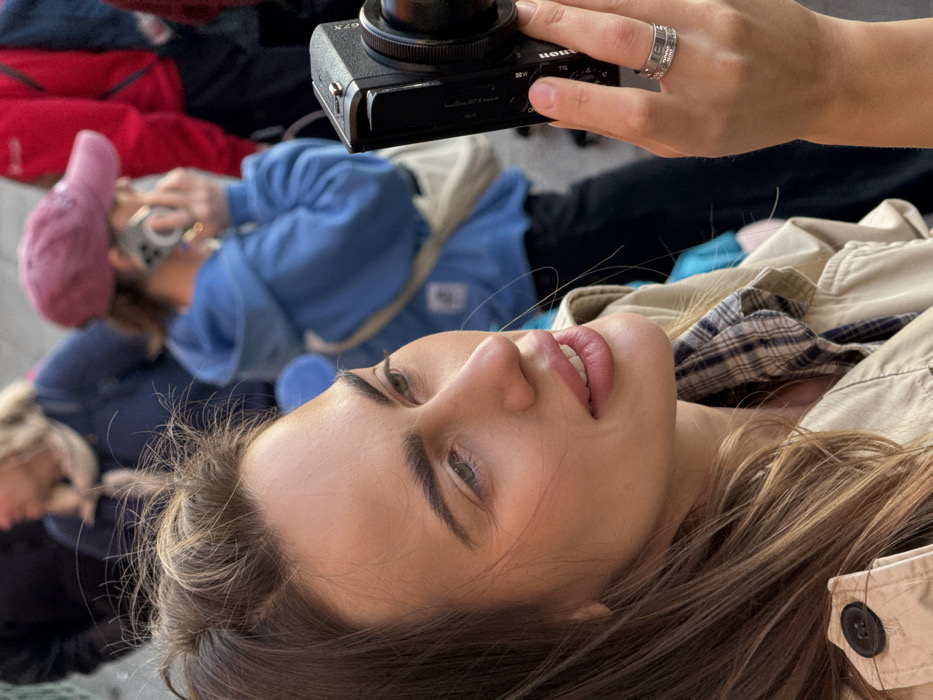 The image presents a close-up, eye-level shot of a female individual in a crowded outdoor urban environment. The specific location within Venice, Italy, is not visually discernible from the available details.
The primary subject is a fair-skinned woman with light brown hair, exhibiting some flyaway strands, looking towards the right side of the frame with a subtle smile. Her eyes are light-colored, and her makeup appears natural, with defined eyebrows and pink-toned lips. She is wearing a light-colored (beige/khaki) trench coat or jacket over a blue and white plaid shirt. Her right hand, prominently featured, holds a black Fujifilm X-T series digital camera with an attached lens; her right index finger rests on a control dial. A silver-toned ring with a textured pattern is present on her right ring finger.
The background is intentionally blurred, depicting several indistinct individuals. In the mid-ground, a person wearing a blue hooded sweatshirt and a pink baseball cap is holding a light-colored smartphone to their right ear, indicating a phone conversation. This individual also wears a light-colored cross-body bag. Additional blurred figures are visible further in the background, including one wearing a dark top and a light scarf, and another in red attire. The scene is illuminated by daylight.