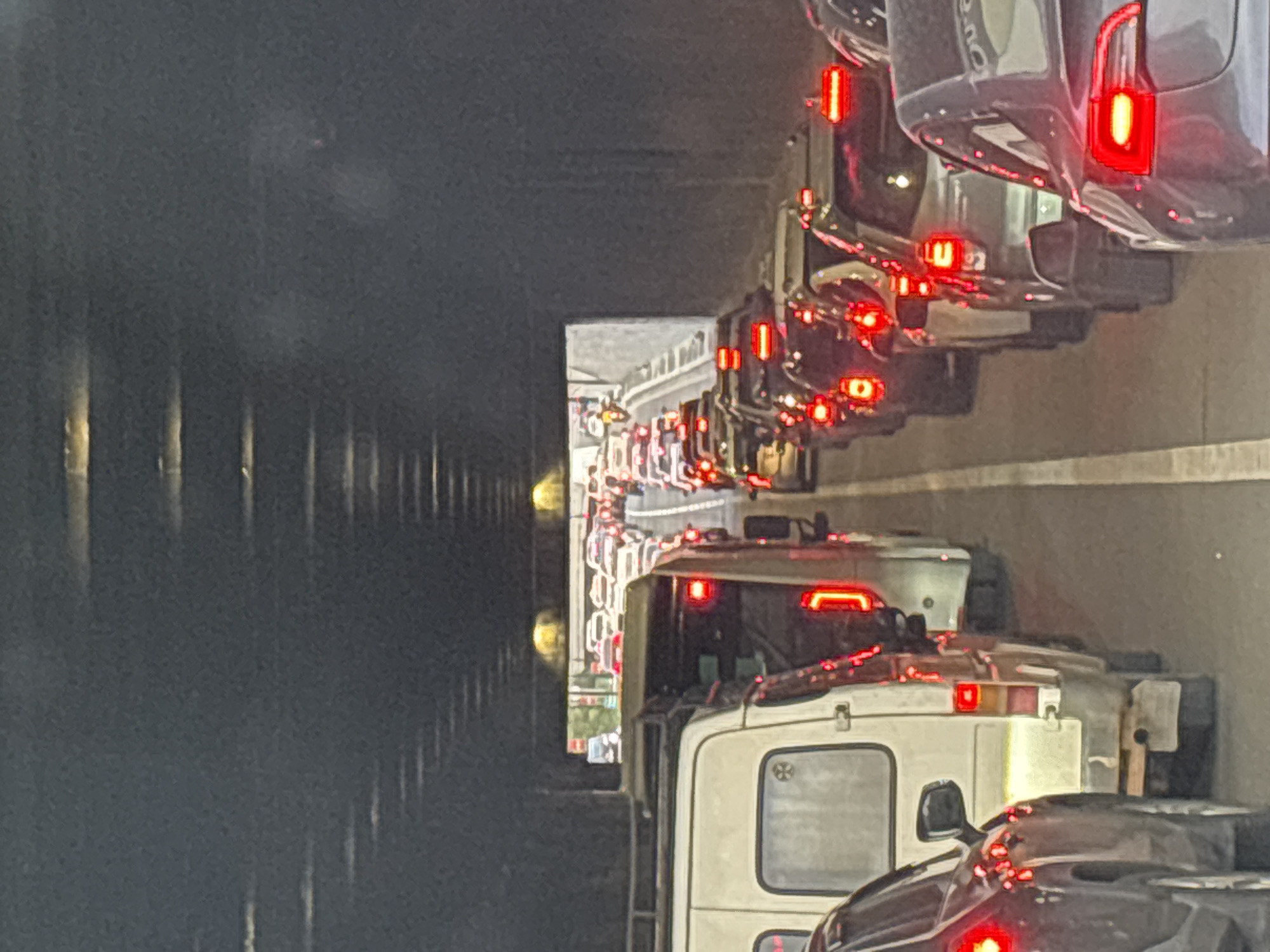 This photograph depicts a severe traffic jam occurring within a dark tunnel or underpass, potentially in Tbilisi, Georgia. The image is captured from a slightly elevated viewpoint, looking down into multiple lanes of stationary or slow-moving vehicles. Countless cars are visible, extending deep into the tunnel, their bright red taillights prominently illuminated.
In the immediate and middle ground, a variety of dark-colored passenger cars are seen alongside a distinct white commercial van, all contributing to the dense congestion. The dark, somewhat reflective walls of the tunnel flank the roadway. At the far end, the tunnel opens up to reveal a glimpse of an urban landscape, possibly including buildings or a multi-level road structure.
The strong contrast between the dark tunnel interior and the glowing red brake lights suggests the time of day is either evening or night. No individuals are clearly visible within the vehicles or on the road, though their presence is implied. The scene is characterized by heavy vehicle density and an apparent halt in traffic flow. No specific text or local identifiers are visible within the image itself to confirm the location.