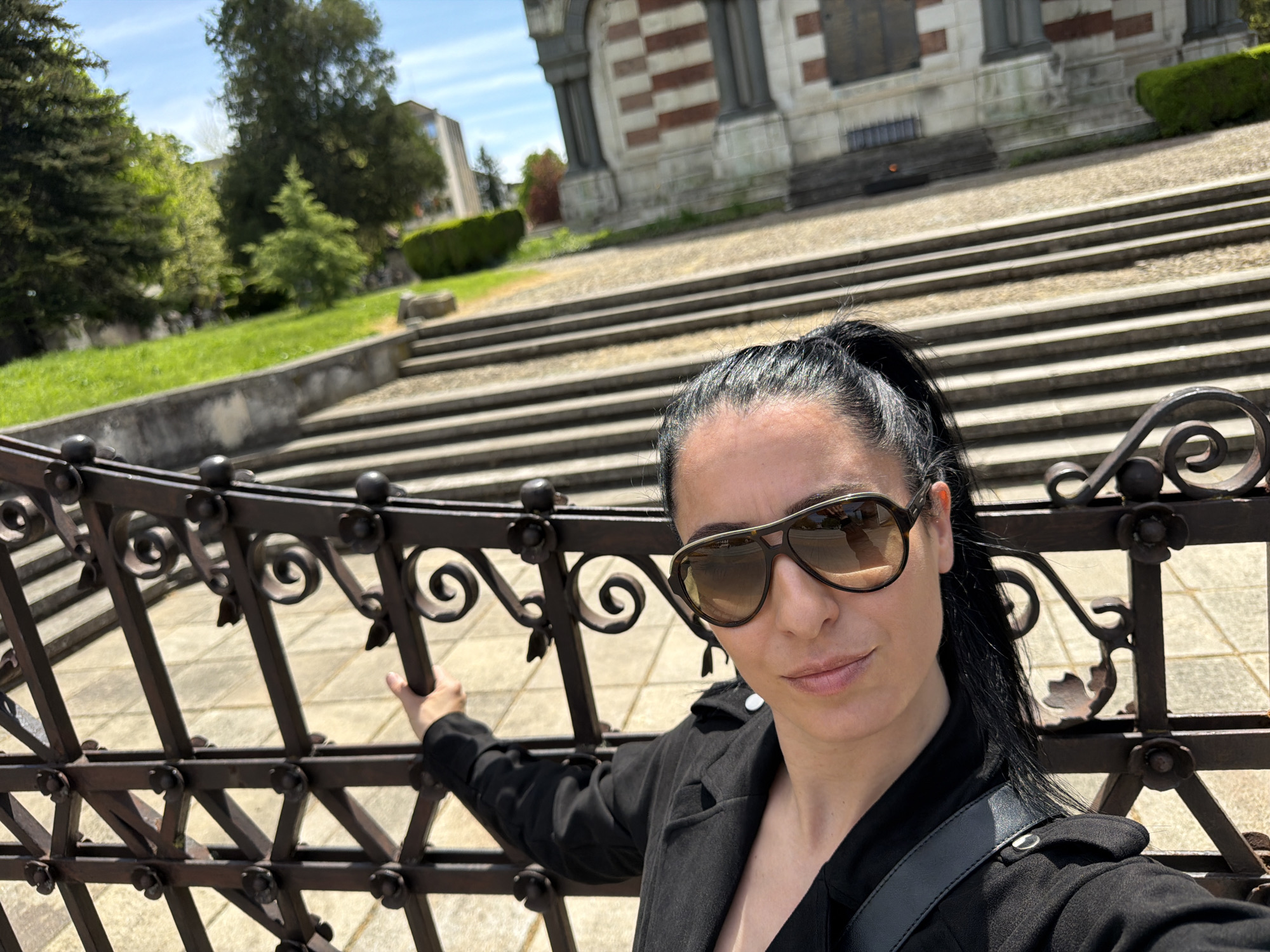 A medium-shot, selfie-style photograph captures a woman with dark hair pulled back in a ponytail. She is wearing large sunglasses and a black jacket, holding onto a dark metal fence. In the background, there are many stairs.
The location is near the Pleven Panorama in Pleven, Bulgaria. A large building with a patterned, brick facade is visible at the top of the stairs. Trees and greenery can be seen to the side of the stairs. The scene is bathed in sunlight, suggesting it is daytime.