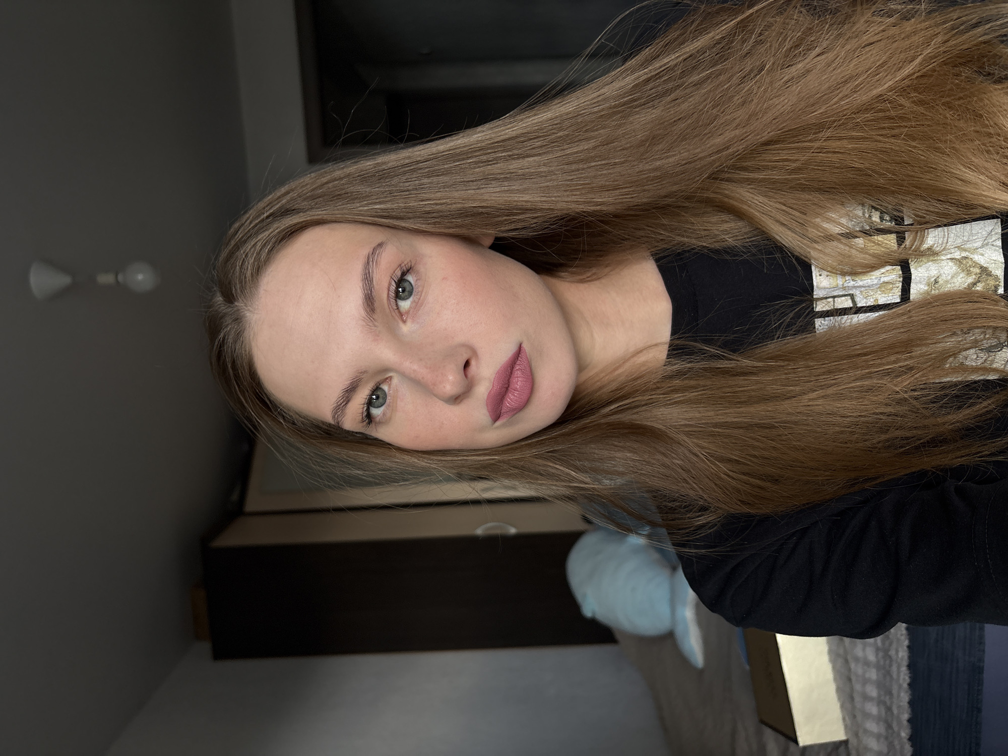 A young woman with long, light brown hair is taking a selfie in a room that appears to be a bedroom. She is wearing a black t-shirt. The background shows a closet, a bed with a light blue stuffed toy, and a small box on the bed. The lighting is dim, with a ceiling light visible in the upper part of the image. There is no indication that this location is in the city of Perm, Russia.