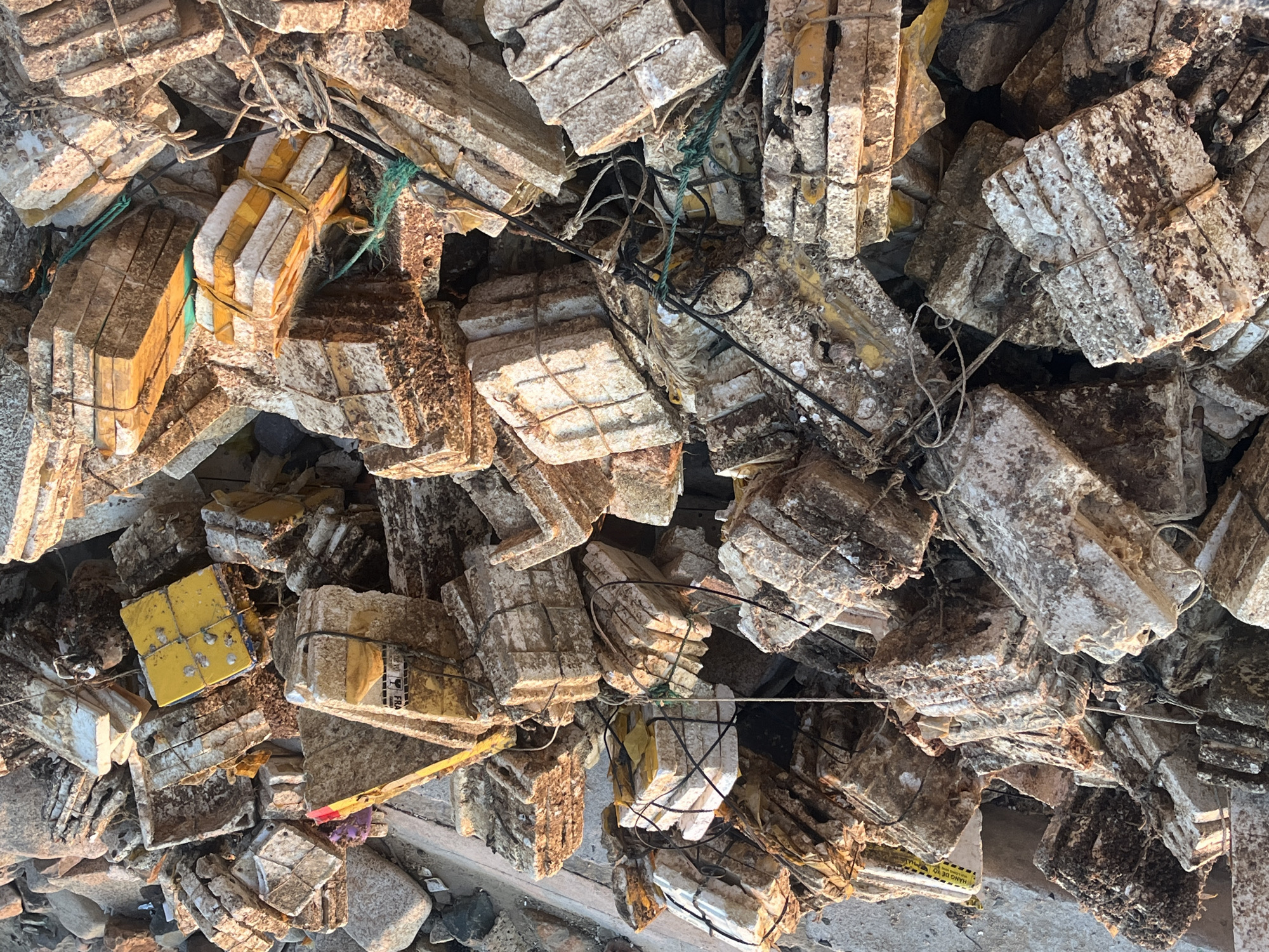 The image shows a large pile of rectangular, block-like objects, many of which appear to be stacked and bound together with string or tape. The objects have a weathered, textured surface, with colors ranging from light tan to dark brown. Some appear to have been partially submerged, judging by the coloration. Several pieces of string and tape are visible among the blocks, indicating a binding or packaging process. The location is likely a coastal environment in Bac Nha Trang Ward, Vietnam, with the items possibly being related to aquaculture, construction, or marine activities. There are no people visible in the image. The scene suggests an accumulation or disposal of these objects.