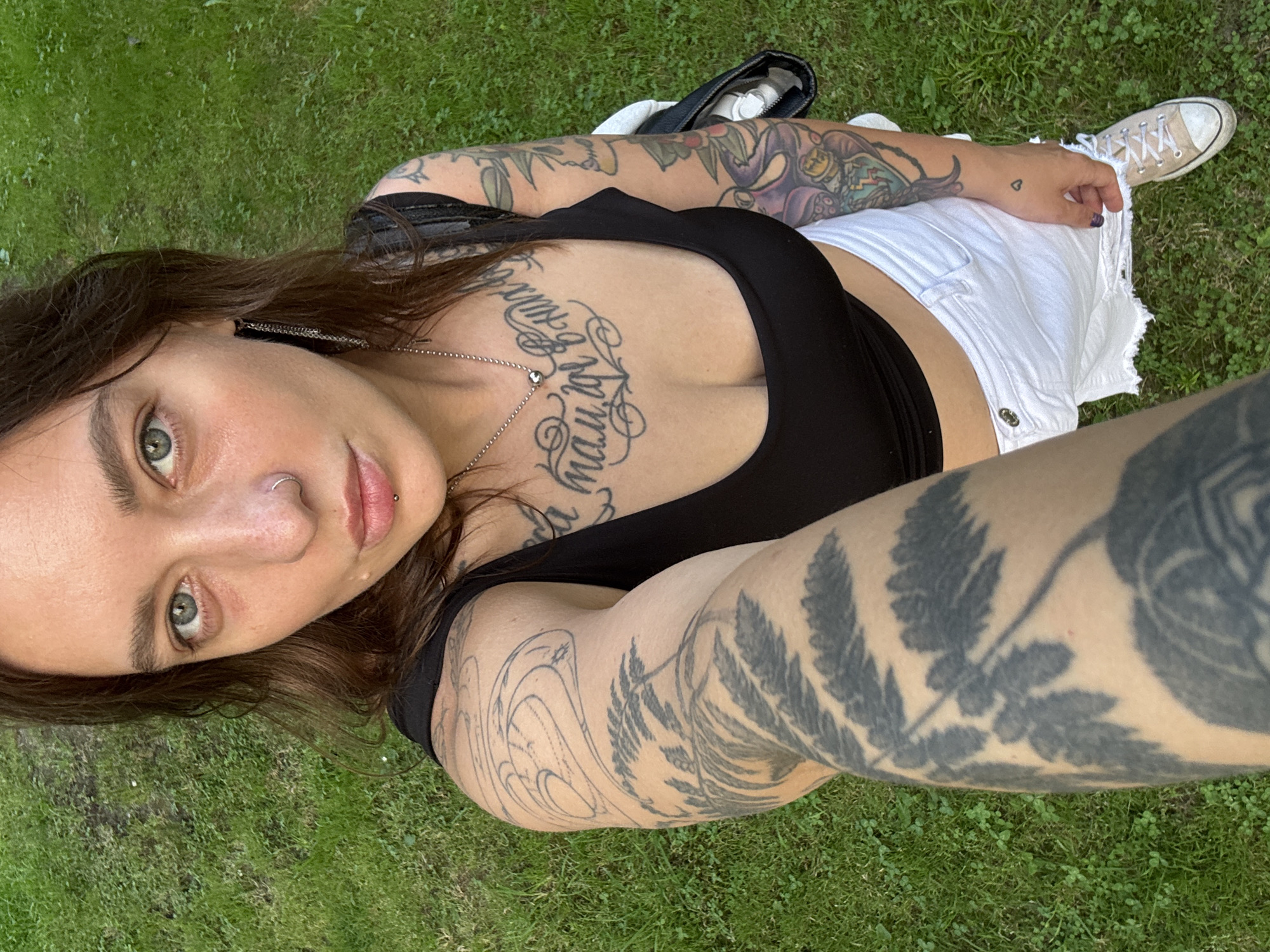 A person is standing in a grassy area, possibly a park or yard, with green grass covering the ground. The person is wearing a black tank top, white distressed denim shorts, and light-colored sneakers. They have numerous tattoos on their arms and chest. The person has a nose ring and a lip piercing. Their left arm is extended towards the camera, showing a large tattoo of fern leaves. The person is looking directly at the camera with a neutral expression. The lighting suggests it is daytime. There are no identifiable landmarks or clues that would place this image specifically in Sochi, Russia.