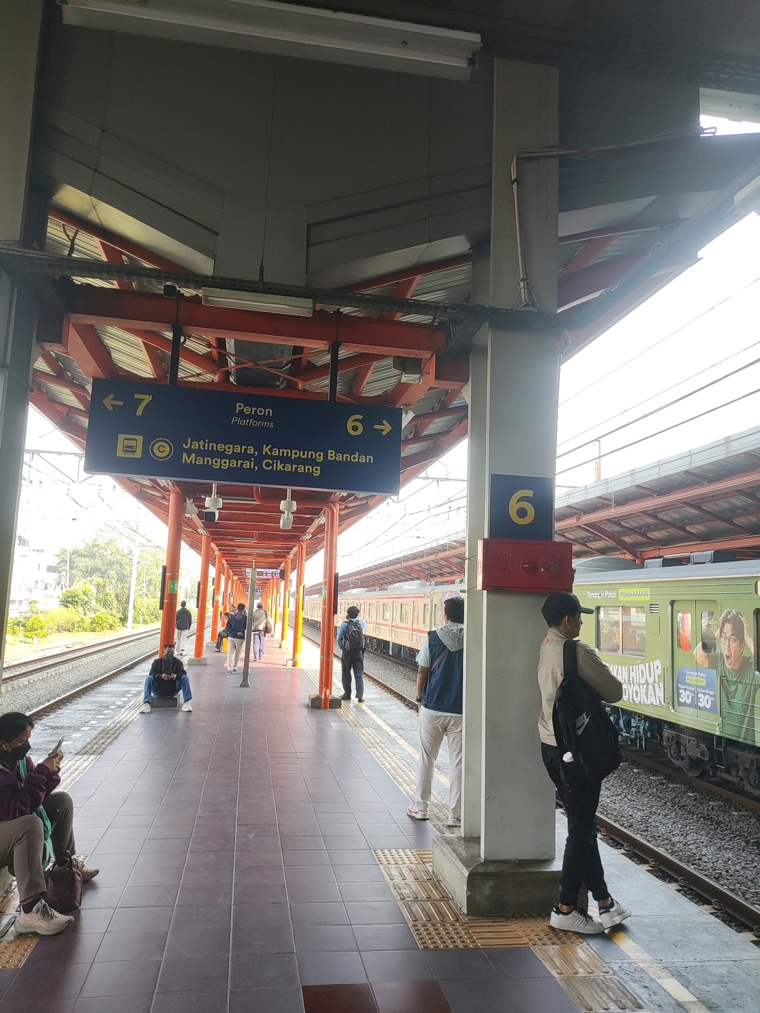 The image depicts a train station platform in Bekasi, Indonesia. Several individuals are visible on the platform, some seated, others standing. A train is present on the tracks alongside the platform. Signage indicates platform numbers and destinations. The environment appears to be an open-air, covered walkway with a tiled floor.