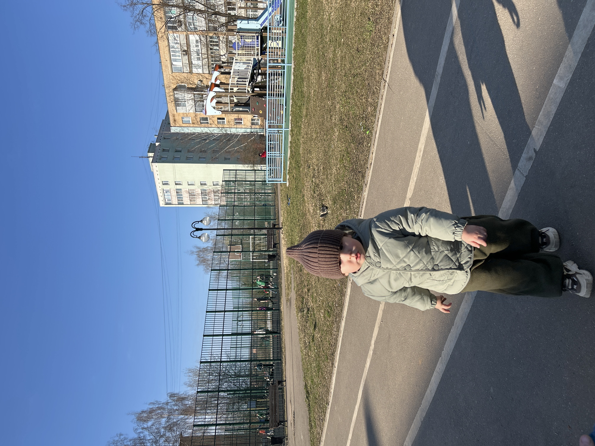 A child wearing a brown knit hat, a quilted light green jacket, and dark green pants is lying on a gray asphalt surface with white lines. The child's face is visible and appears to be looking at the camera. In the background, there is a green mesh fence, bare trees, and a clear blue sky. Behind the fence, there is a playground structure and two multi-story buildings. The scene is in daylight. The location is likely the city of Klin, Russia, based on the provided information.