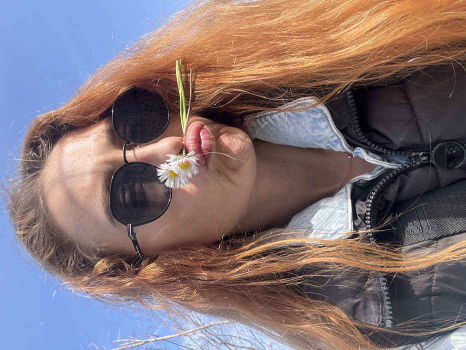 A close-up outdoor portrait depicts a female subject under a clear blue sky. The individual has long, reddish-brown hair and wears dark, round sunglasses. Her lips are puckered in a pout, holding two white daisies with yellow centers and green stems between her upper lip and nose. She is dressed in a light blue denim collar visible beneath a dark, zippered padded vest. A thin chain necklace with a small pendant is visible on her neck. The background consists of a bright, unclouded blue sky, with indistinct bare tree branches visible on the left side of the frame. No specific landmarks are present to identify a precise location within Budva, Montenegro.