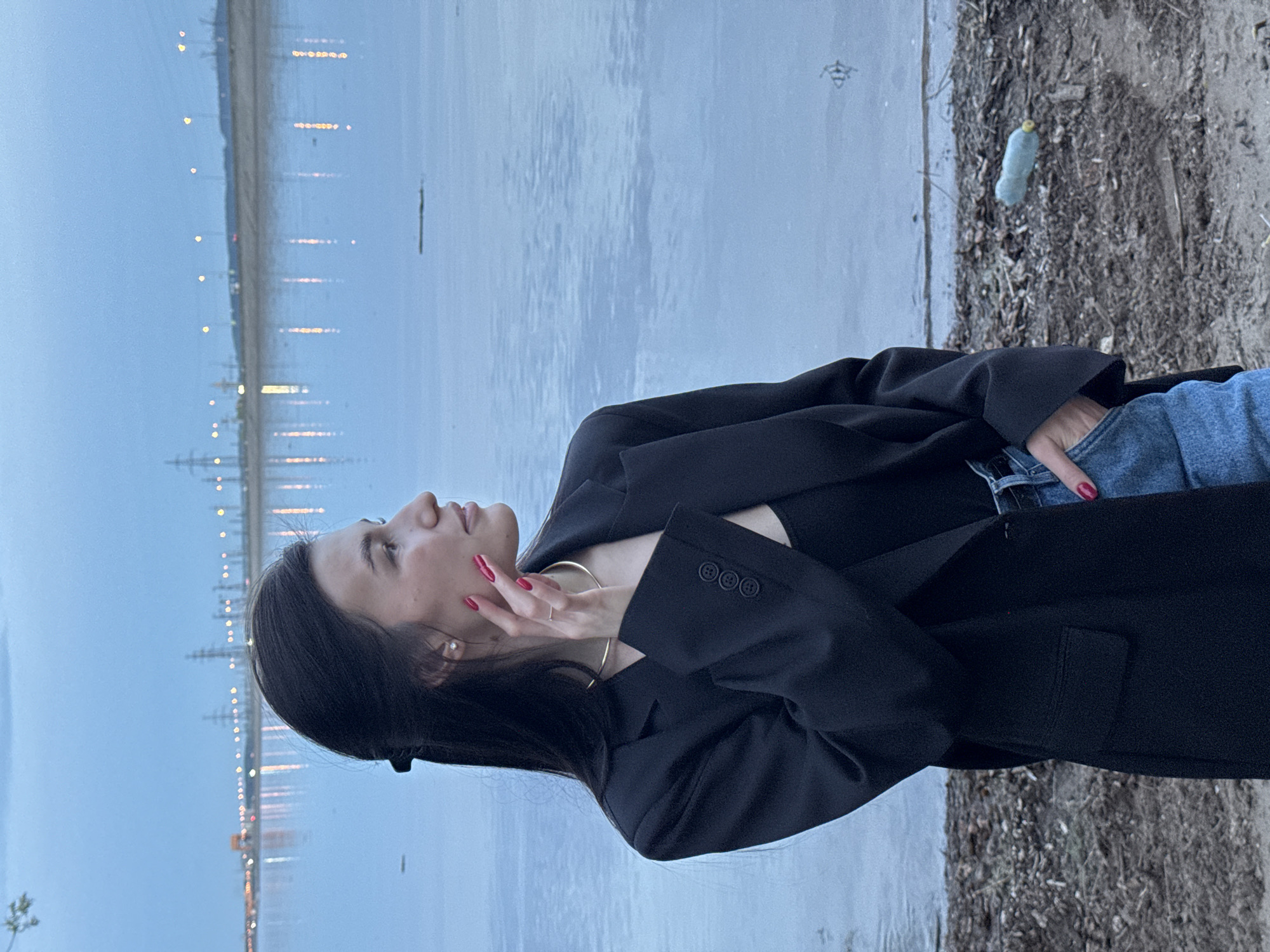 A woman stands on a bank, likely near the Kama River in Chaikovsky, Russia, given the cityscape across the water. She is wearing a black blazer over what appears to be a black top, and blue jeans. Her right hand is raised to her face. The background features a body of water with light reflections from city lights across the water and sparse vegetation or debris on the near bank.