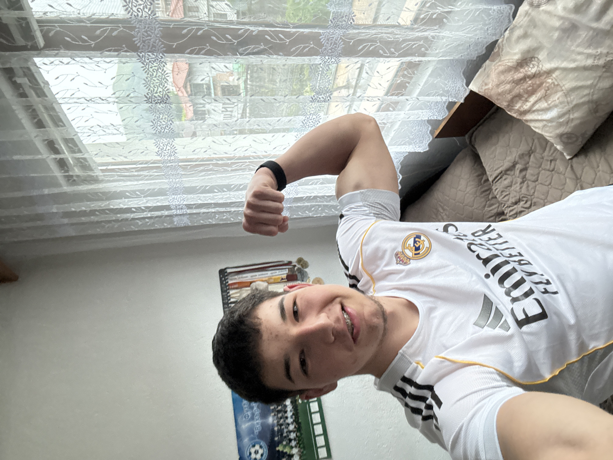 A young man takes a close-up selfie indoors, flexing his left arm and smiling cheerfully, revealing braces on his upper teeth. He is wearing a white Real Madrid football jersey with the Adidas logo and "Emirates Fly Better" sponsor text visible on the front. A black wristband is on his left wrist.
The setting is a brightly lit room, likely a bedroom or living area in Zlatograd, Bulgaria. To the man's right, a window is covered by sheer white curtains featuring a distinct patterned design. Through the curtains, glimpses of green foliage and distant buildings suggest an outdoor, possibly residential, scene under daylight. On his left, a dark shelf containing books or binders is partially visible, along with a poster depicting a football field. Behind him and to his right, a sofa or bed is covered with several textured and patterned pillows in neutral tones. The man's expression conveys happiness and playfulness.