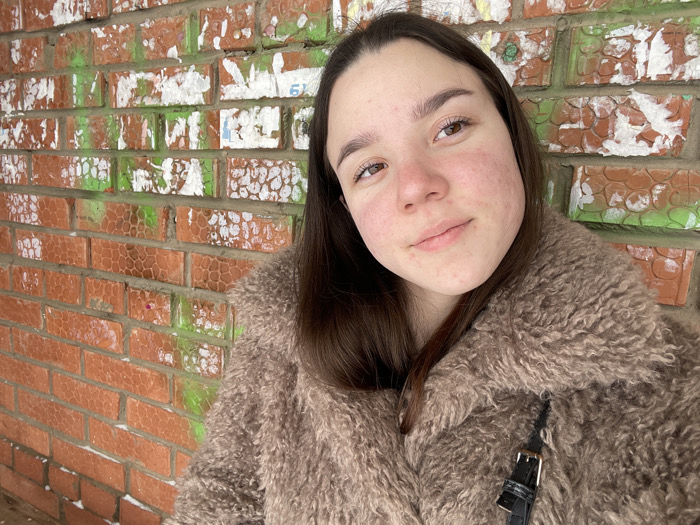 A portrait-oriented photograph captures a young female subject positioned against a brick wall. The location in the city of Ольховка, Russia, cannot be confirmed from the visual evidence. The female, characterized by medium-long dark brown hair, fair skin with noticeable redness on the cheeks and nose, and light brown eyes, looks directly at the camera with a subtle, closed-mouth smile. She is dressed in a thick, textured, light brown faux fur coat. A black strap with a gold-colored buckle, indicative of a shoulder bag, is visible over her right shoulder/chest area. The background consists of a reddish-orange brick wall with a distinct textured pattern on the bricks. This wall is heavily marked with irregular, peeling patches of white material, likely remnants of torn posters or paint, and substantial splashes of bright green paint. The lighting is soft and diffused, suggesting an overcast outdoor environment.