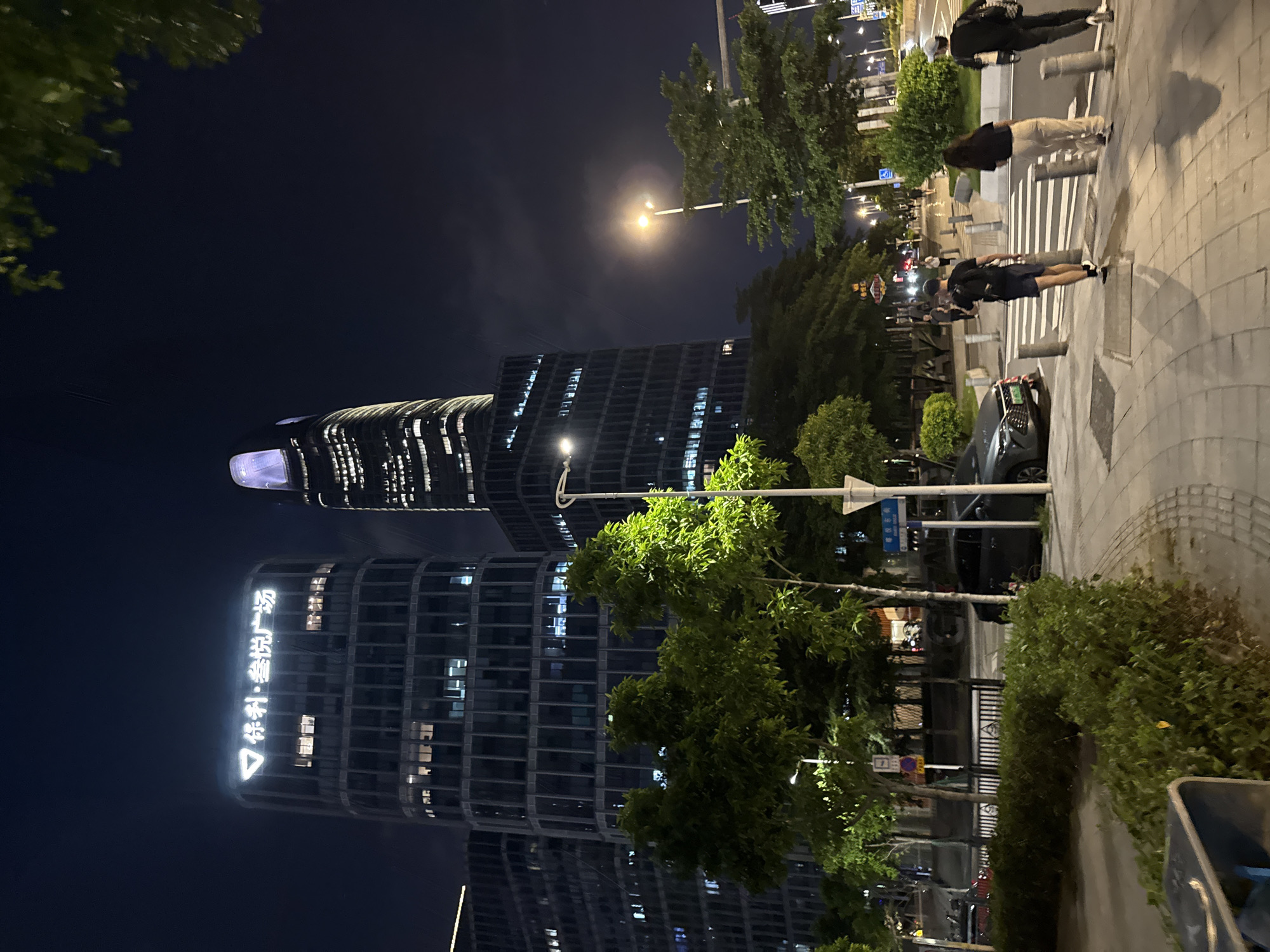 This media file captures an urban street scene at night in Haizhu District, China. The dominant subjects are two modern, illuminated skyscrapers with distinctive curved designs. One building displays prominent white text, possibly a company logo or name, in Chinese characters. A streetlamp casts a warm glow on the scene, creating a slightly hazy atmosphere, suggesting a clear night.
Several people are visible walking along a paved pedestrian area. They appear to be casually strolling, with one individual holding a phone, perhaps taking a photo or video. The presence of a parked dark-colored car and street signs with Chinese characters further confirms the location. Lush green trees line the sidewalk, adding a touch of nature to the urban environment. The overall mood is tranquil, characteristic of a city winding down for the evening. The time of day is clearly night, indicated by the darkness of the sky and the artificial lighting illuminating the buildings and street.