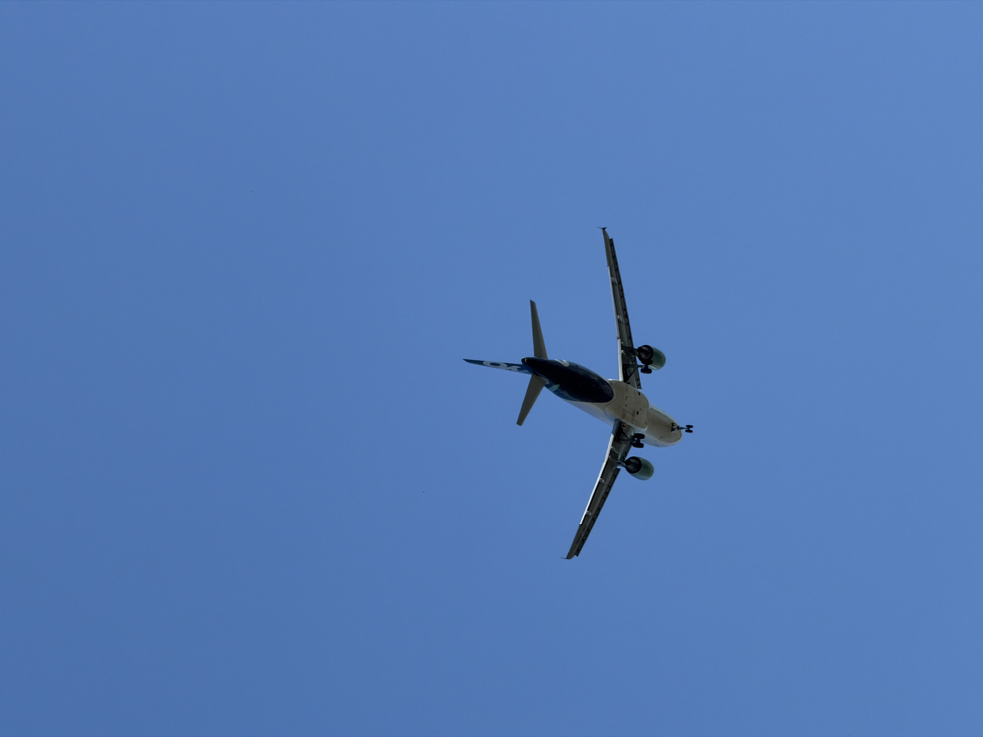 An airplane is flying in a clear blue sky, seen from below. The underside of the aircraft is visible, showing its wings, engines, and fuselage. The tail of the plane features a dark blue color with a logo. The landing gear is extended, suggesting the plane is either approaching or departing from an airport. The background is a uniform expanse of sky, devoid of any clouds or other visible features. There is no indication of the city of Burgas, Bulgaria, or any specific location within it in this image.