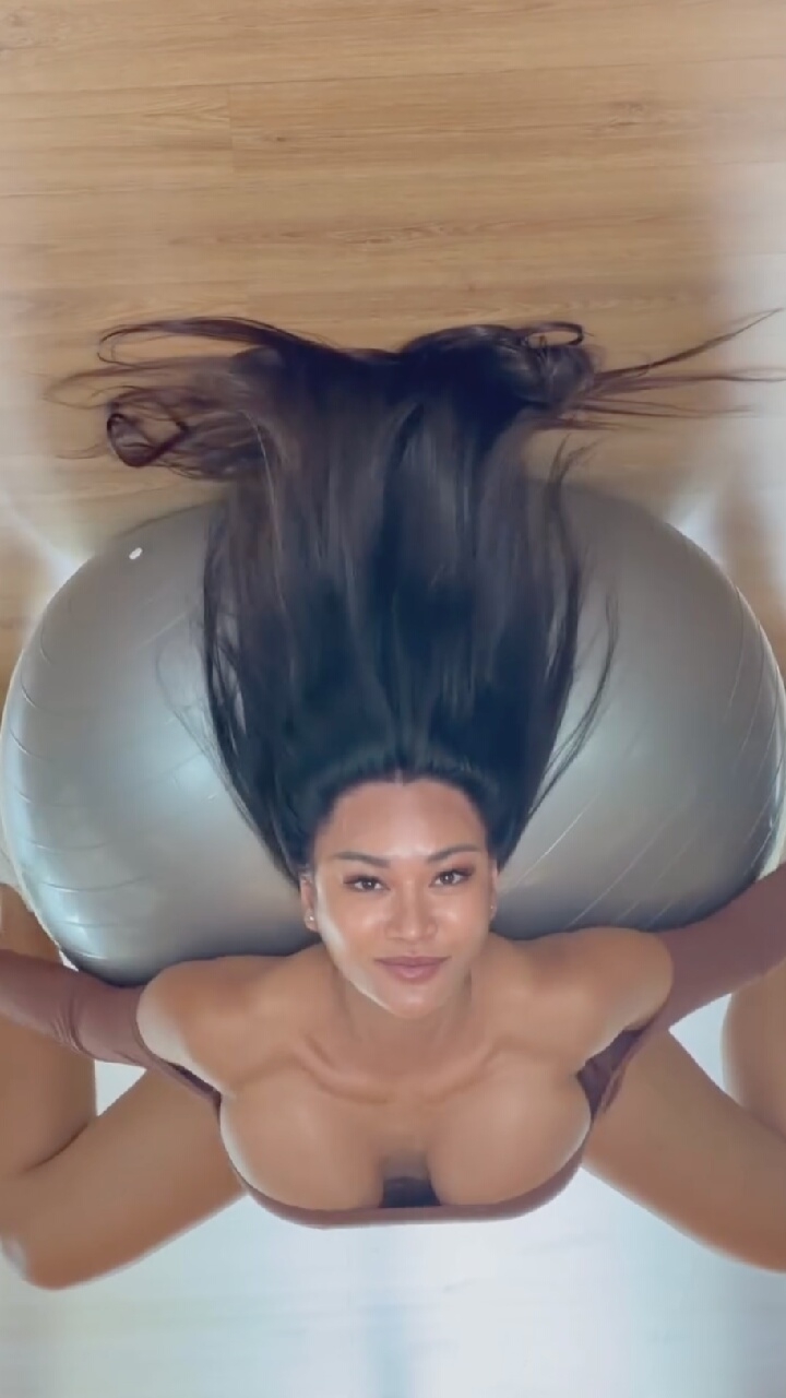 A woman with long dark hair is lying on a wooden floor, supported by a large exercise ball. She is wearing a brown top and her arms are extended outwards. Her legs are spread apart. The image is taken from a high angle, looking down at the woman. There is no indication of the location being Moscow, Russia.