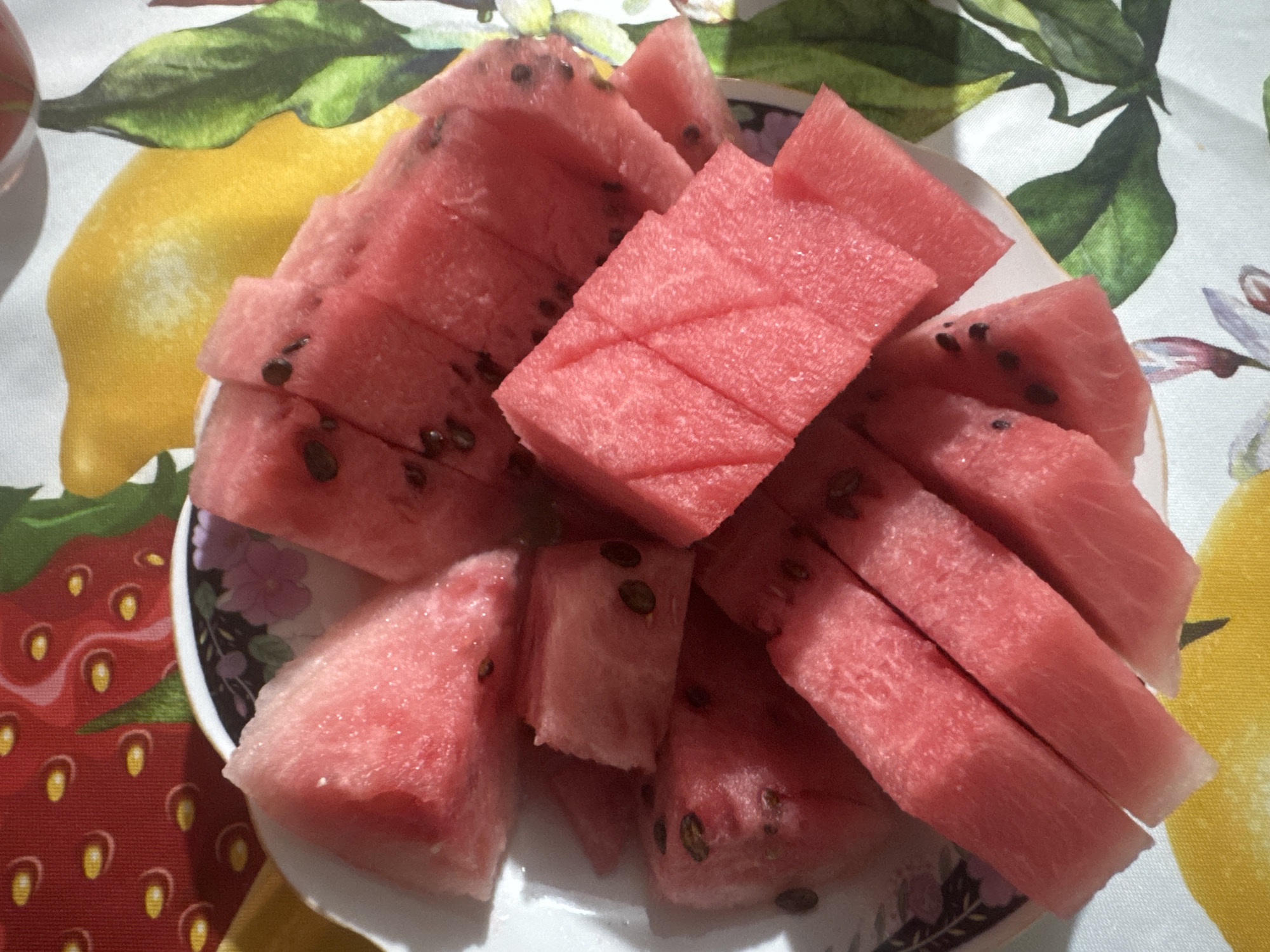 A close-up, overhead shot shows a white plate filled with sliced watermelon, positioned on a colorful, patterned tablecloth. The tablecloth features a design of strawberries, lemons, and green leaves. The watermelon slices are a vibrant pink, with visible seeds scattered throughout. Some slices are arranged in neat stacks, while others are scattered more casually on the plate. The lighting suggests an indoor setting. There is no indication of the city of Chardara, Uzbekistan in the image.