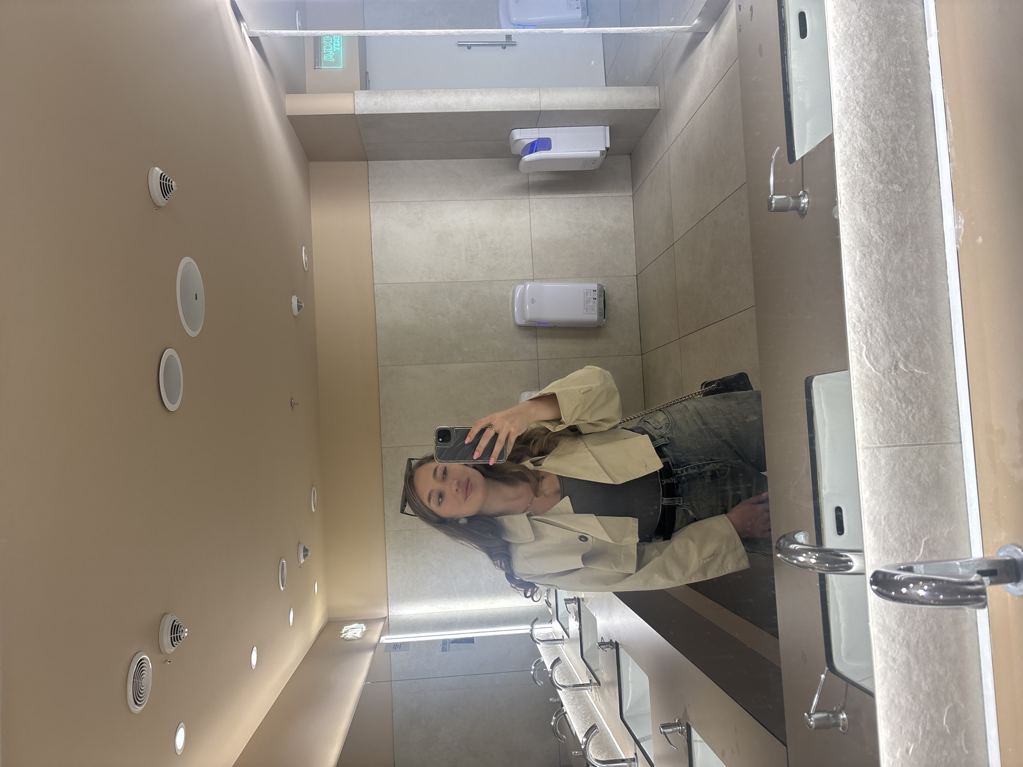 A young woman is taking a selfie in a public restroom. She is wearing a beige jacket, a gray tank top, and jeans. She has sunglasses on her head. The restroom has multiple sinks with stainless steel faucets and white countertops. There are automatic hand dryers and soap dispensers on the wall. The ceiling is off-white with round vents and speaker-like fixtures. The walls are tiled in a neutral color. A green exit sign is visible near the top of the image. The location is likely in Kharkiv, Ukraine, as indicated by the prompt.