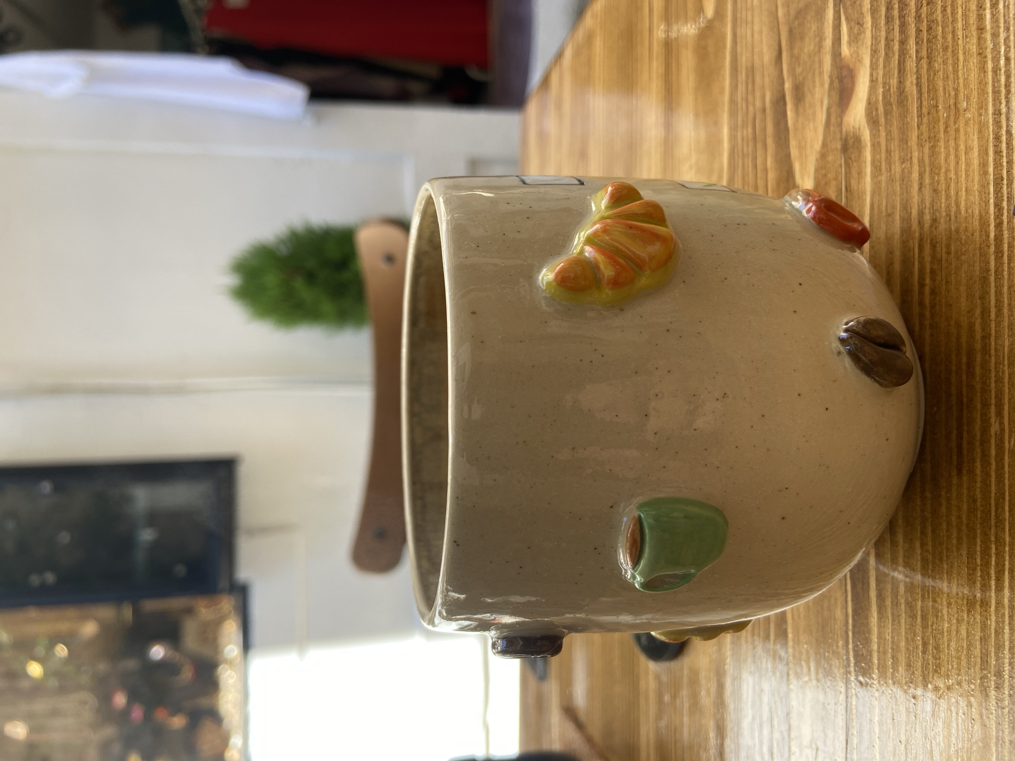 A close-up, eye-level shot captures a beige ceramic mug adorned with miniature, three-dimensional food items. The mug features a coffee cup, a croissant, coffee beans, and a red chili pepper. It rests on a polished wooden table. In the blurred background, a small green plant is visible, along with hints of a light-colored wall and a dark framed object.