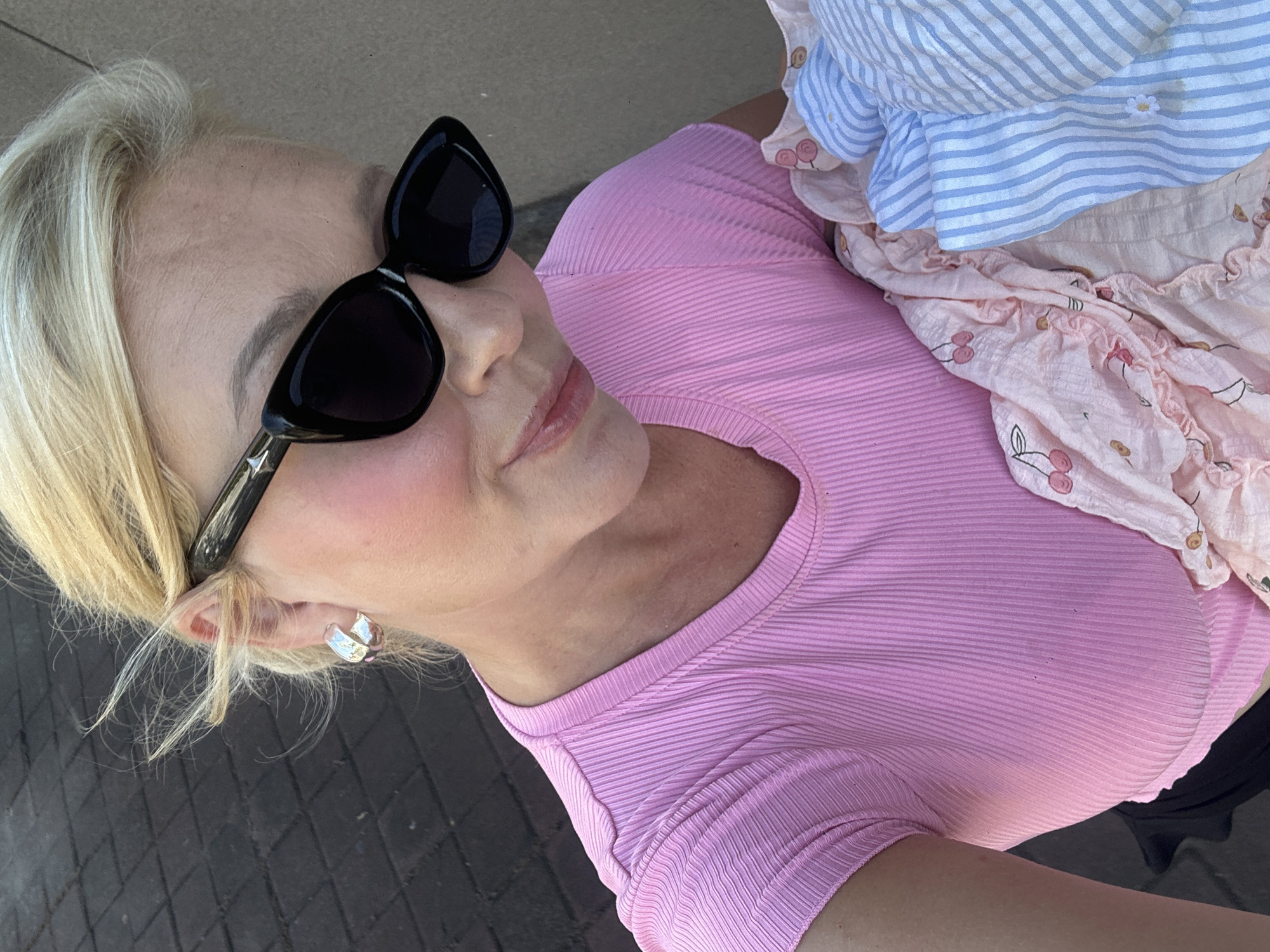 A woman stands on a brick surface, possibly a walkway or sidewalk. She is wearing black sunglasses with a star detail on the frame, a pink ribbed top, and silver earrings. She is holding a light-colored patterned garment, possibly clothing, with blue and white stripes and a cherry design. The location may be North Kuta, Indonesia.