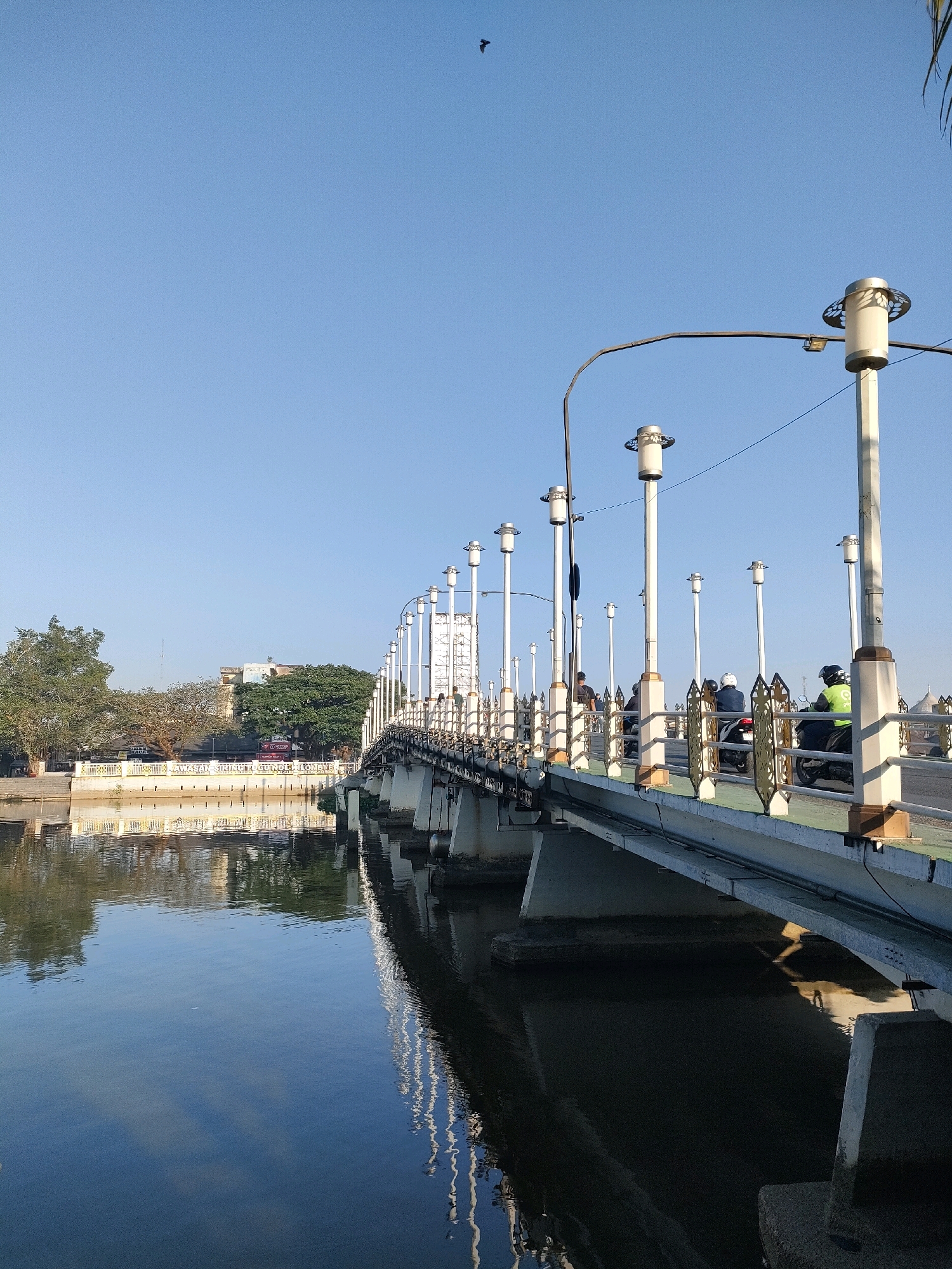 This image captures a view of a bridge in the city of Banjarmasin, Indonesia, likely the Jembatan Banuaanyar. The bridge spans a calm body of water, with its concrete pillars and superstructure reflected in the dark water. The bridge is lined with numerous decorative lampposts, each topped with a white cylindrical light fixture.
On the bridge, several people are visible. Two individuals are riding motorcycles, one wearing a bright green safety vest. Another person is walking on the bridge, and a few other figures are partially visible in the distance. A metal railing with ornate gold-colored decorative elements runs along the side of the pedestrian walkway.
In the background, across the water, a tree-lined embankment is visible. Buildings are present behind the trees, suggesting an urban or semi-urban setting. The sky is a clear, bright blue, with a single bird in flight high above. The overall lighting suggests it is daytime, likely morning or late afternoon due to the quality of light.