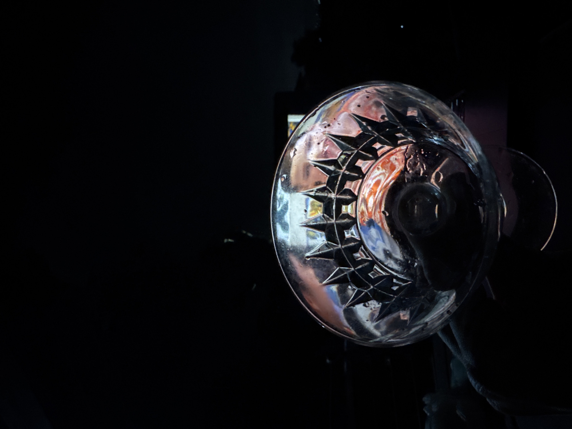 A close-up shot, taken from a low angle, reveals a crystal glass with a starburst design. The glass is positioned on the right side of the frame against a dark background. The bowl of the glass has a distinctive shape, and the design consists of repeating starburst patterns around its circumference. Reflected in the glass are hints of colors and shapes, suggesting an interior setting. The photograph's background is almost entirely black, creating a sense of depth and emphasizing the light reflecting off the glass. The location is not discernible from the image. No people are visible.