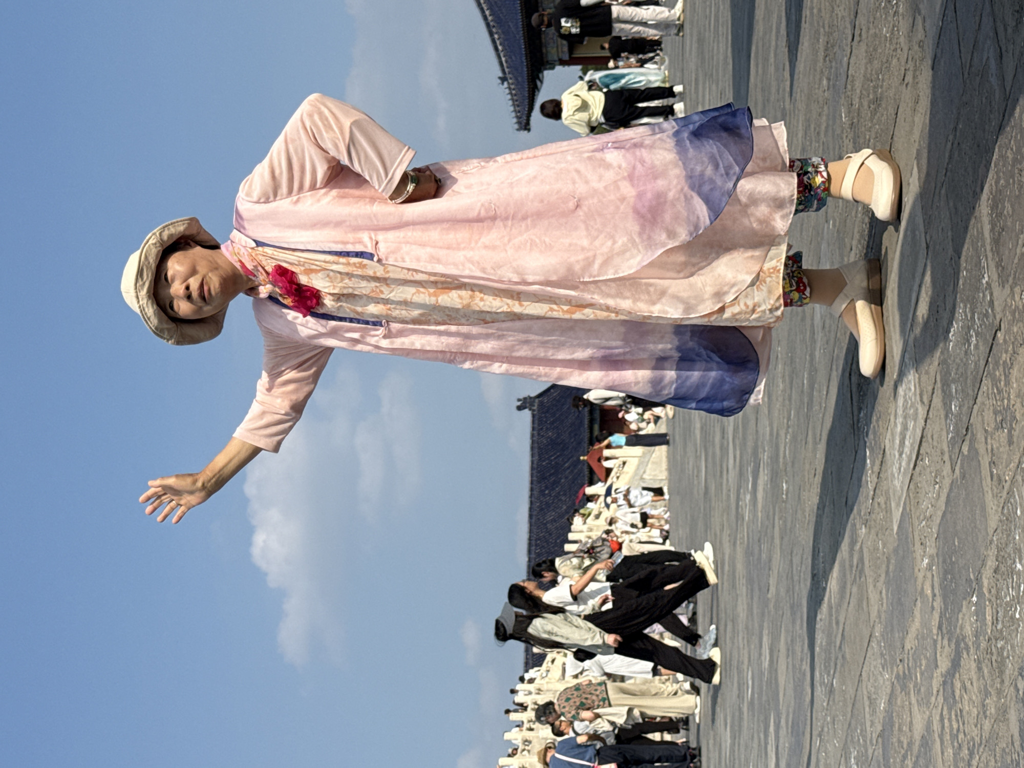 An elderly Asian woman wearing a light-colored sun hat, a long light pink garment with a floral pattern on the inner layer, and colorful patterned pants is standing in the foreground of the image. She is waving with her right hand, which is raised. She is also wearing light-colored slip-on shoes. Behind her, there is a stone courtyard with several people walking. The background features traditional Chinese architecture with dark blue tiled roofs and white decorative elements. The sky is clear and blue with a few scattered clouds. The overall scene appears to be a tourist area in the Dongcheng District of China.