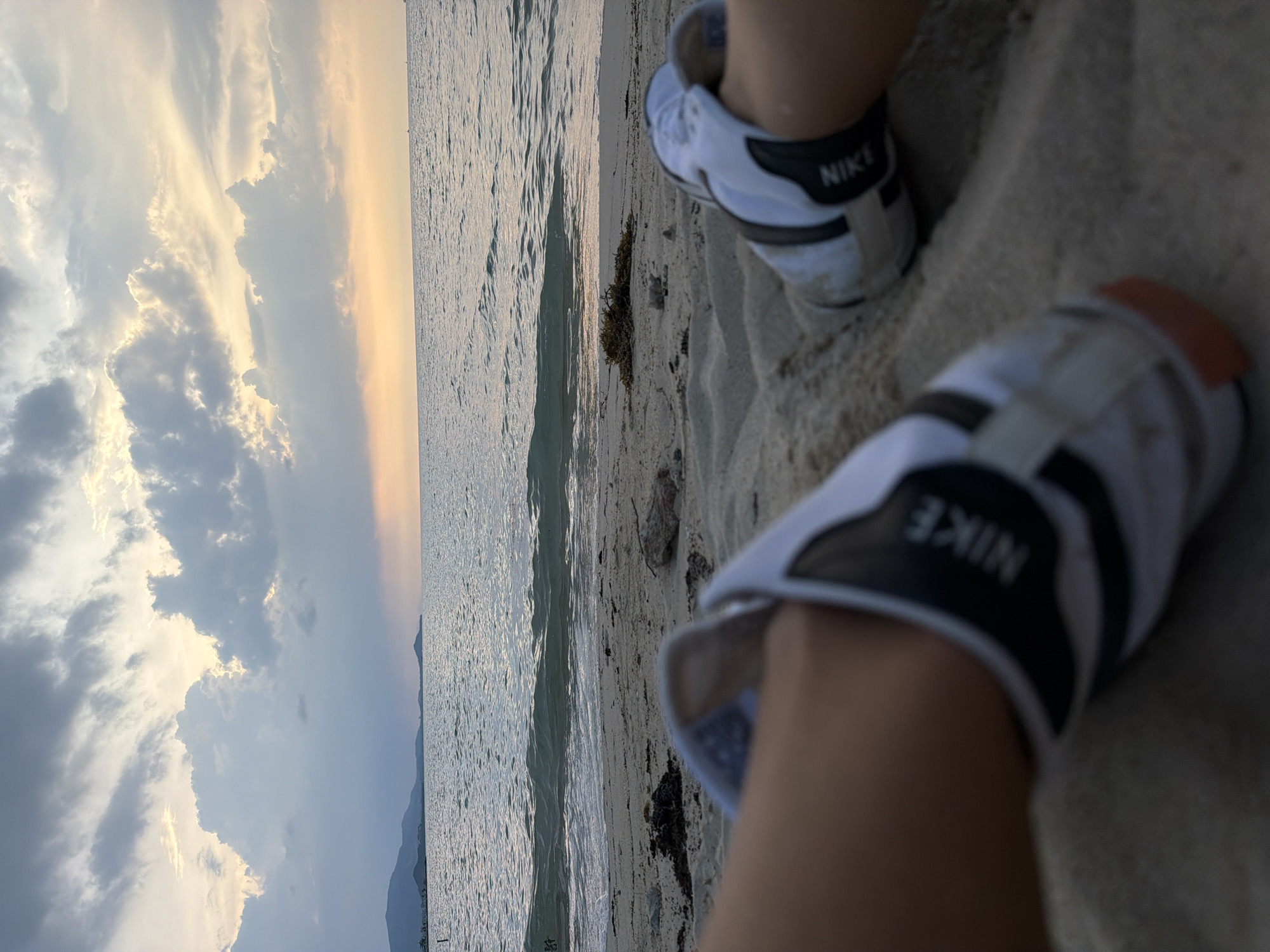 A first-person perspective shot shows a person's feet, wearing white Nike sneakers with black accents, partially submerged in sand on a beach. The sand is a light tan color and appears to be fine-grained. In the background, the ocean stretches out to the horizon, with gentle waves lapping at the shore. The sky above is filled with dramatic clouds, transitioning from a deep gray on the left to lighter hues and hints of orange and yellow from the setting sun on the right. Distant hills or mountains can be seen along the coastline on the left side of the frame. The overall mood is serene and contemplative, with the natural beauty of the beach and sky dominating the scene. The location is likely Ko Samui District, Thailand, given the tropical appearance of the coastline and the style of the sand and vegetation.