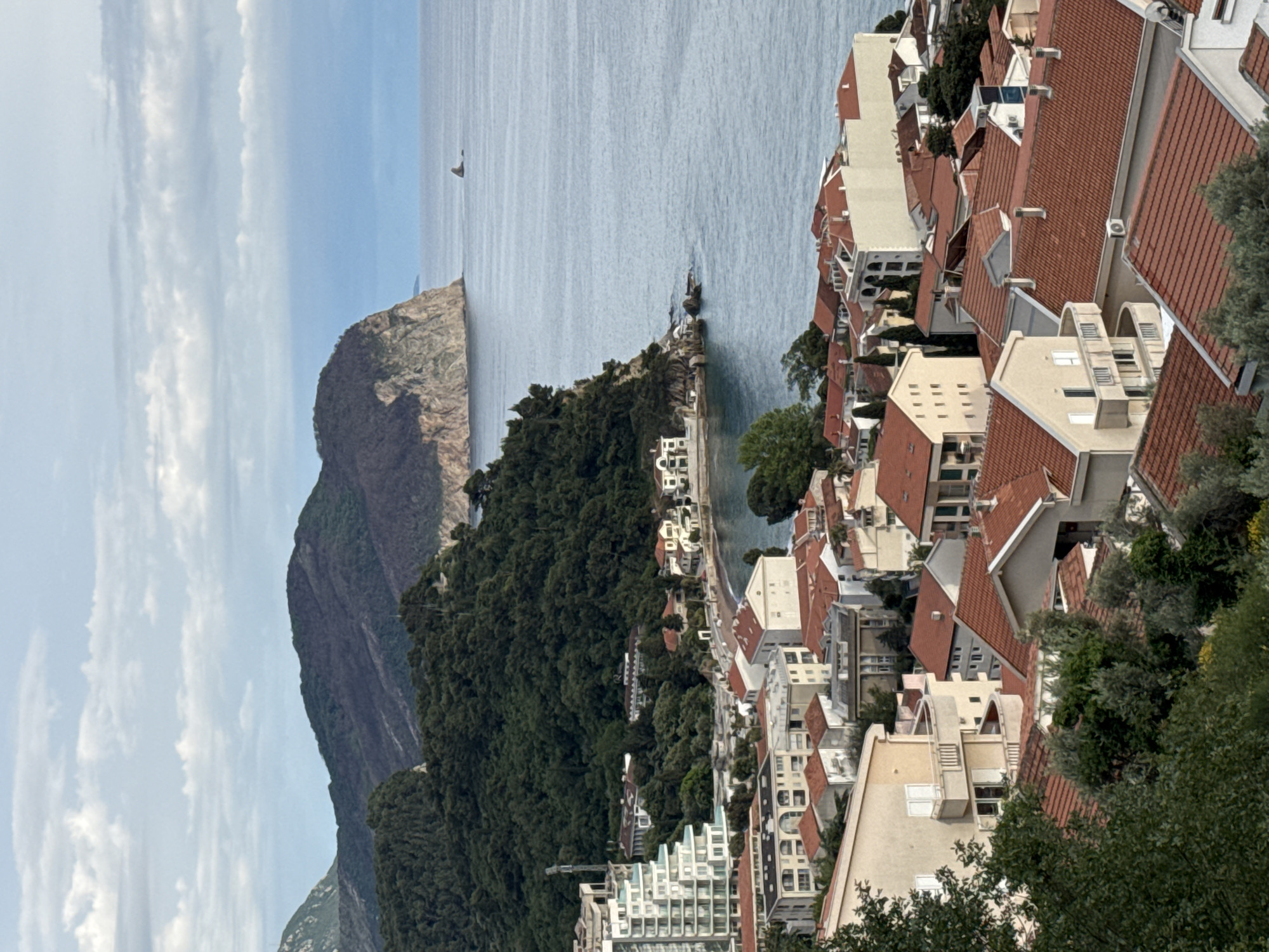 An aerial view depicts a seaside town. To the left, a large, steep mountain rises from the coast. The water is smooth, with a sailboat visible in the distance. On the right, houses with red tiled roofs are clustered together, built on the hillside. The buildings are a mix of residential and commercial structures. Lush green trees are interspersed amongst the buildings, adding a natural element to the urban landscape. The sky is partly cloudy. This is Medinski Krš, Montenegro.