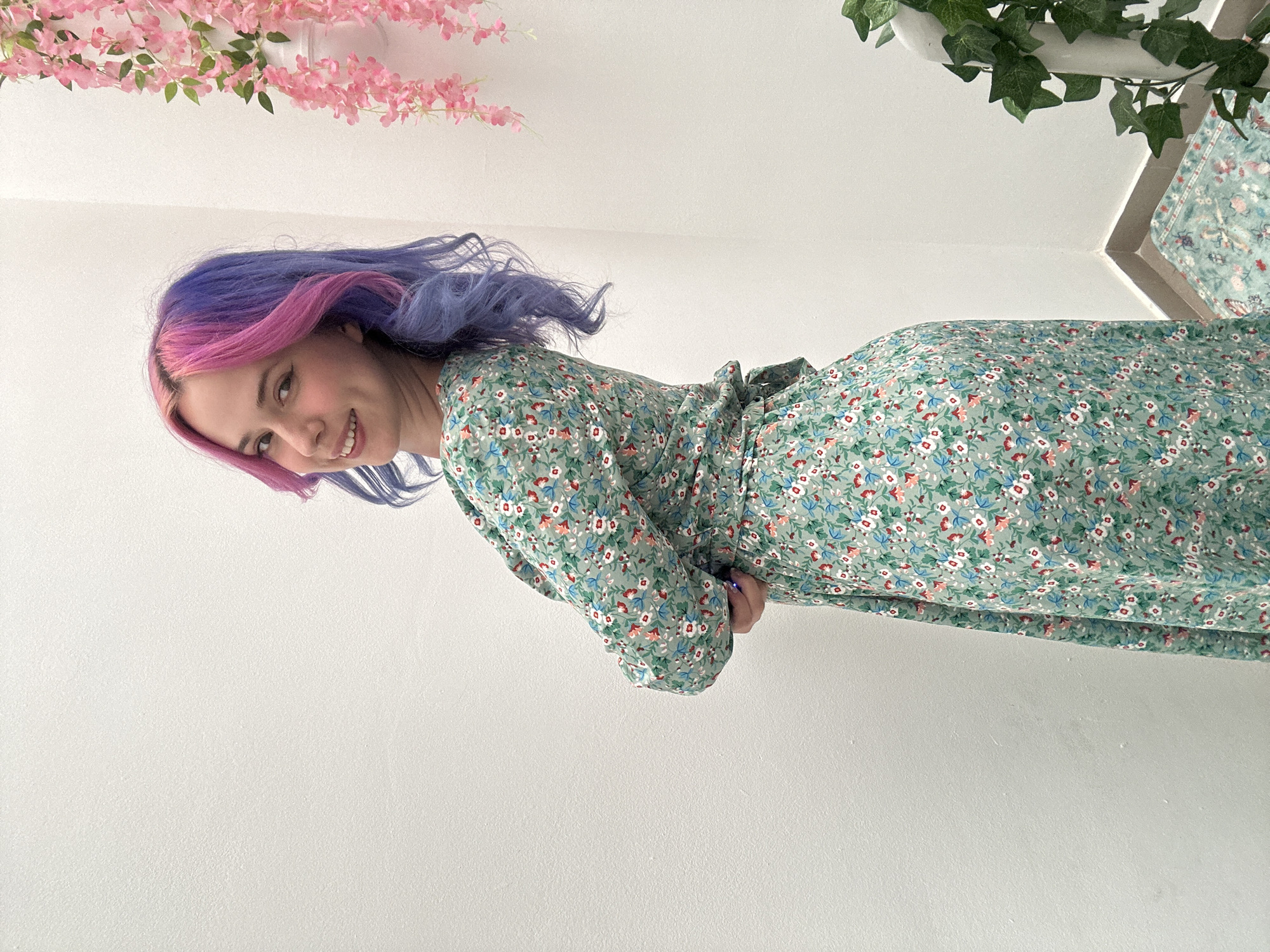 A young woman is depicted in a brightly lit indoor setting, smiling and looking directly at the camera. She has long, wavy hair distinctively dyed in vibrant pink and purple hues. She is wearing a long-sleeved dress with a light green base, adorned with a delicate floral pattern of small white, blue, and reddish-pink flowers with green leaves. The dress features a tie-waist. Her right arm is bent at the elbow, with her hand positioned near her waist.
The background is predominantly clean white walls, suggesting a minimalist interior. Decorative elements include a string of artificial pink cherry blossom-like flowers hanging across the upper left portion of the frame, and artificial green ivy leaves hanging from the upper right, near what appears to be an architectural corner.
The image is presented rotated 90 degrees counter-clockwise, showing the woman horizontally although she is standing upright. The lighting is bright and even, indicative of indoor conditions. The overall mood is cheerful and friendly. No visible text is present in the image. The scene is set in the City of Zagreb, Croatia.