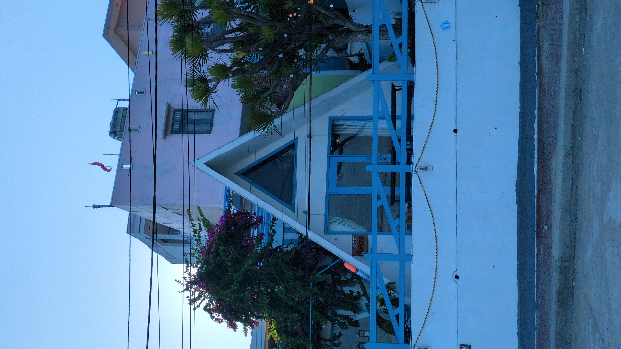 The image displays a multi-storied building with distinctive architecture and vibrant colors, set against a clear, light blue sky. The building appears to be a guesthouse or a small hotel, possibly located in a warm, coastal, or Mediterranean region.
The upper section of the building, occupying the left side of the frame, is painted a soft pink or faded magenta. It features several rectangular windows with dark frames. On its flat roof, a dark-colored water tank is visible, alongside two small red flags or banners that appear to be fluttering gently.
The lower and more prominent section of the building, closer to the viewer and occupying the right side, features a white facade. A central part of this section boasts a distinctive triangular roofline. Within this white area, there are striking blue-framed elements: a large triangular window on the left, and a rectangular door or window unit to its right. A bright blue wooden railing with diagonal cross-braces extends across the front of this white facade, adding a decorative and structural element. Strung horizontally along the white wall, just above the railing, are two decorative ropes secured by hooks. A small, orange rectangular sign with the number "8008" written in black is attached near the blue-framed door/window. A small blue circular emblem with an indiscernible design is also present on the white wall above the ropes.
Lush vegetation enhances the scene. A large tree, possibly a palm or a similar tropical species, with green fronds, grows from behind the blue railing and extends upwards, partially obscuring the pink upper story. Small decorative lights, resembling fairy lights, are strung through its branches. Below this, a vibrant bush adorned with purple or magenta bougainvillea flowers cascades against the white wall, providing a rich burst of color.
Numerous black utility lines crisscross the upper left portion of the image, stretching against the clear sky. The ground level, visible at the bottom right of the image, shows a paved surface, likely a sidewalk or a narrow road, with varying textures of grey concrete and reddish-brown earth.
The clear sky and soft, directional lighting suggest either early morning or late afternoon, casting gentle shadows and highlighting the building's colors. The overall atmosphere is bright and inviting, typical of a lively, warm climate.