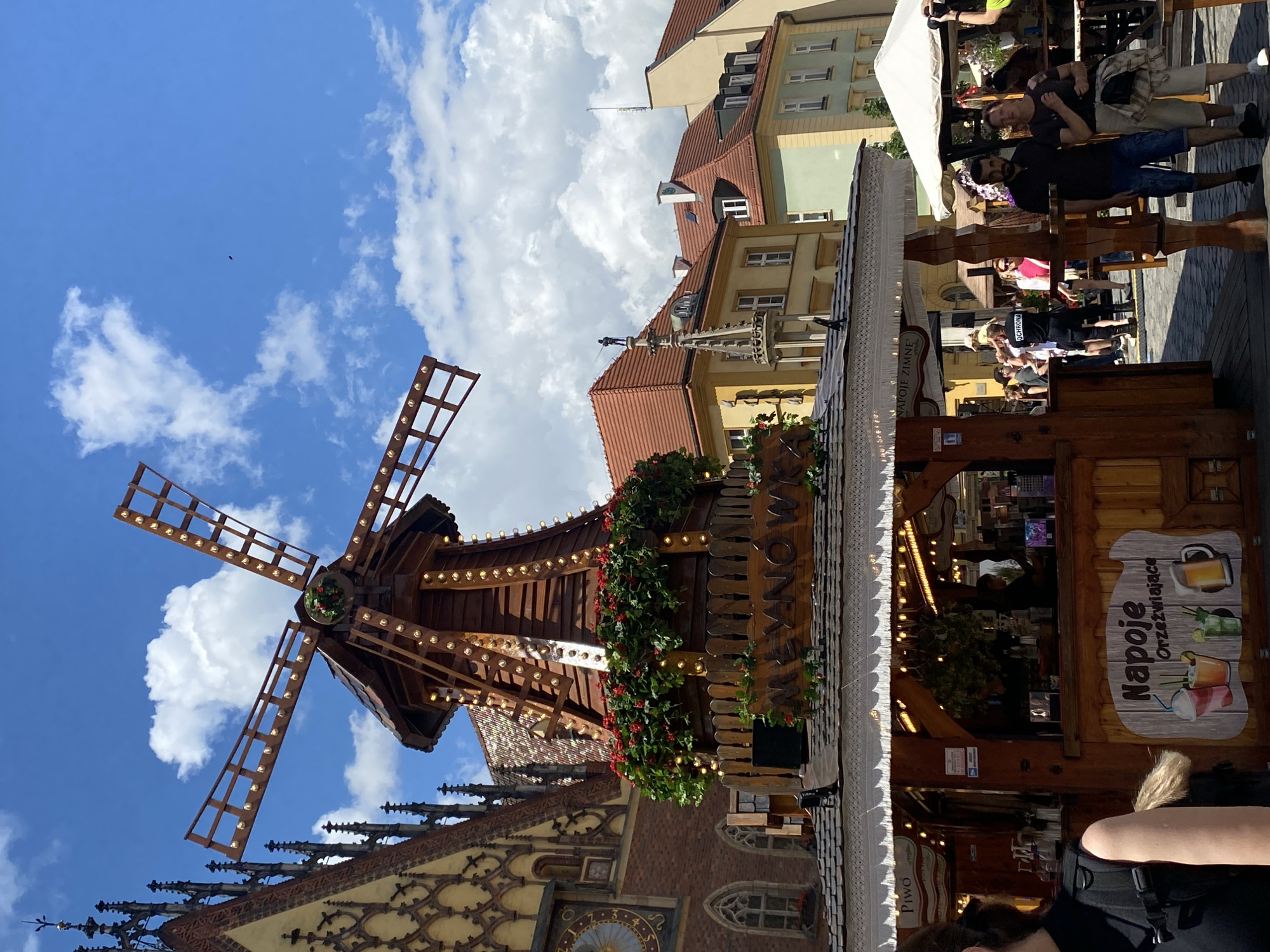 This image depicts a bustling outdoor scene in Wrocław, Poland, specifically in the Market Square (Rynek). The central and dominant feature is a large, elaborate wooden windmill structure, functioning as a decorative element and likely part of a market stall. The windmill is dark brown, adorned with numerous yellow lights outlining its blades and body, and a circular band of red flowers and green foliage around its mid-section. A wooden sign on its base reads "MŁYNÓW" (Młynówka).
Beneath the windmill is a wide market stall with a dark, shingled roof and a white, decorative frill along its edge. The stall itself is made of light brown wood. On the left side of the stall, a sign is partially visible, reading "PIWO" (beer). Towards the right, a large wooden sign on the stall's front displays "Napoje Orzeźwiające" (Refreshing Drinks) with illustrations of various beverages like a cocktail, a mint drink, and a beer mug. Inside the stall, several individuals are working behind a counter.
In the foreground on the left, the back and left shoulder of a person wearing a black sleeveless top and a backpack are visible.
The background features several historical buildings under a bright blue sky with scattered white clouds. To the left of the windmill, a large gothic-style building, likely the Old Town Hall, is visible, characterized by intricate brickwork, pointed spires, and a large golden clock face with Roman numerals. Further to the right, behind the windmill, are other multi-story buildings with red-tiled roofs, varying in color from light yellow to pale green. One building features a slender, ornate spire topped with a dark, small statue of a person, possibly a fisherman. A small, dark, elongated object, possibly a bird or a drone, is visible high in the sky to the right of the windmill's blades.
Numerous people are visible throughout the scene, walking, standing, and congregating around the market stalls. On the right, a white market tent is present. One person in the mid-ground wears a black t-shirt with "OCHRONA" (Security) written on the back. The ground is paved with cobblestones. The lighting indicates a bright, sunny day.