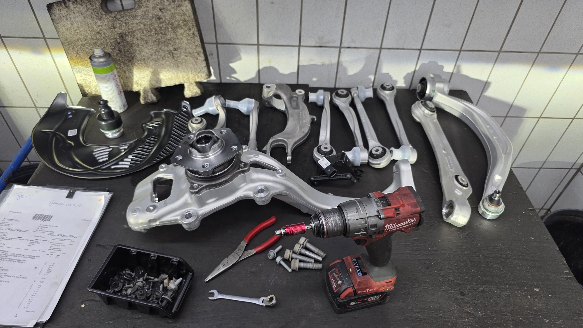 In an indoor workshop setting, likely a garage, a collection of car suspension components and tools are laid out on a dark surface. The objects include several metallic control arms of various shapes and sizes, a wheel hub assembly, a brake dust shield, and a black plastic container filled with bolts and screws. A red Milwaukee cordless drill is positioned prominently, with a driver bit inserted into a bolt. Red-handled pliers and a small wrench are also visible. A partially visible document, possibly an invoice or repair order, is situated to the left of the components. The background features a tiled wall and a rough, textured mat with a spray can on top. The lighting suggests overhead illumination casting shadows on the tiled wall. There are no people present in the image.