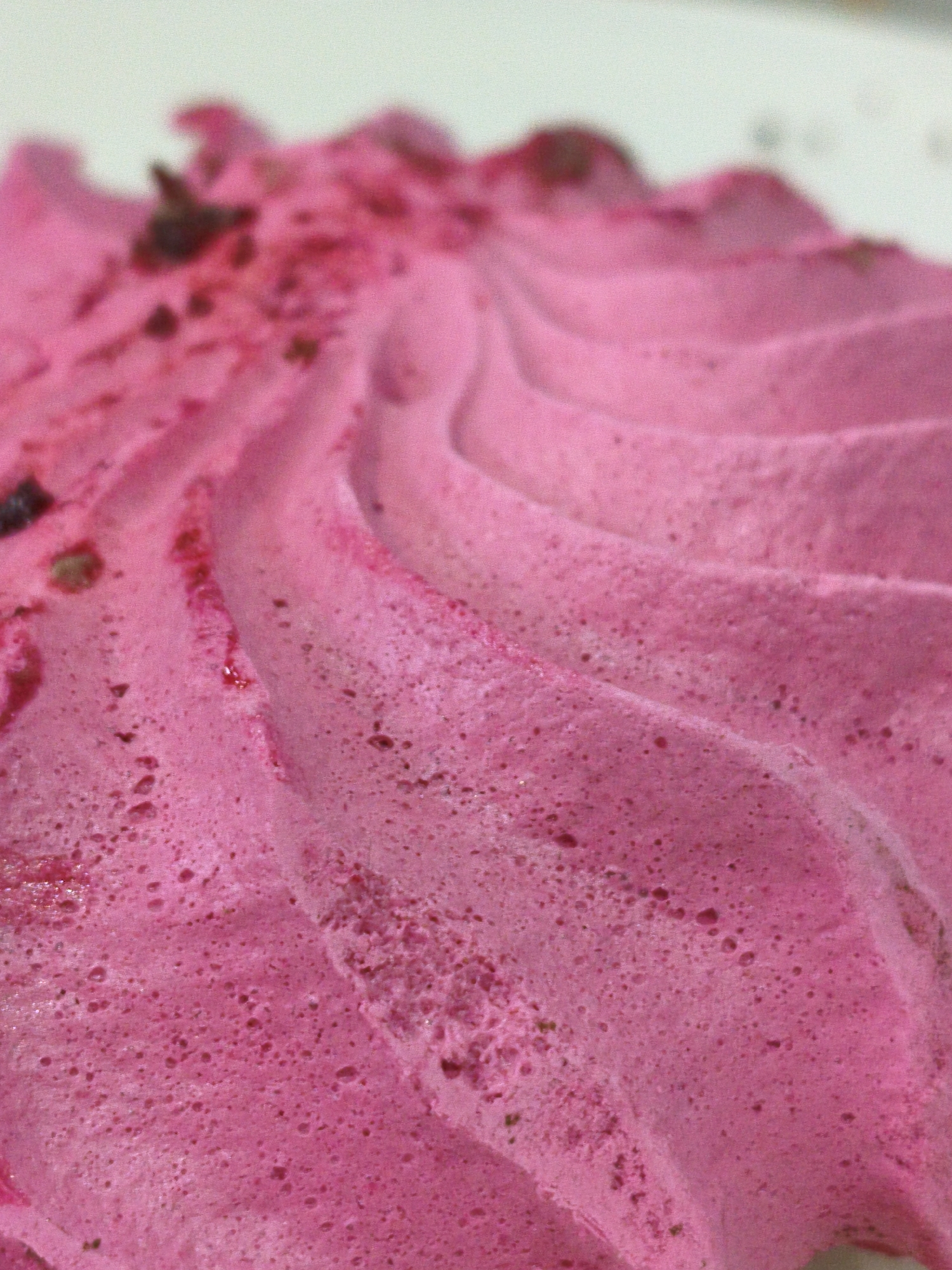 The image shows a close-up, macro shot of a pink, foamy dessert, likely a mousse or whipped cream, with visible air bubbles and small, darker specks embedded within. The dessert is swirled, creating a textured, wave-like pattern. The background is out of focus and appears to be a plain, light surface. There are no people, animals, or any elements that would suggest a specific location in Orenburg, Russia, or any other geographical area. The focus is solely on the texture and color of the dessert.