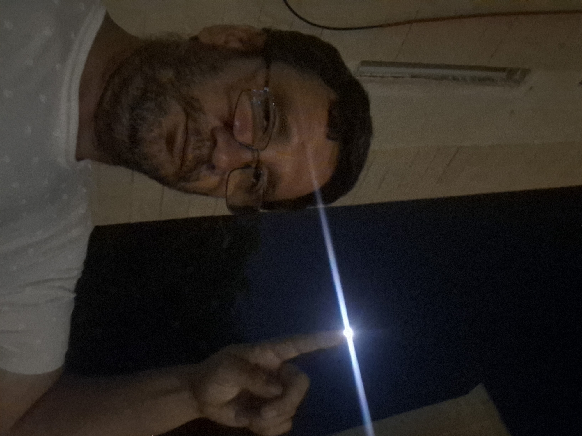 A close-up, medium shot captures a man with short, dark hair, glasses, and a beard. He is wearing a white t-shirt and is looking towards the camera. His right hand is raised, with his index finger pointing upwards, seemingly towards the moon, which is obscured by a bright flare in the night sky. The background consists of a dark, blurry expanse, possibly indicating a night setting outdoors. The man is positioned in the foreground, while the moon and the dark background occupy the upper right portion of the frame. The lower left of the image shows a pale wall with horizontal lines. The overall impression is of a selfie taken at night, with the man interacting with the night sky. This image was taken in Yerocham, Israel.