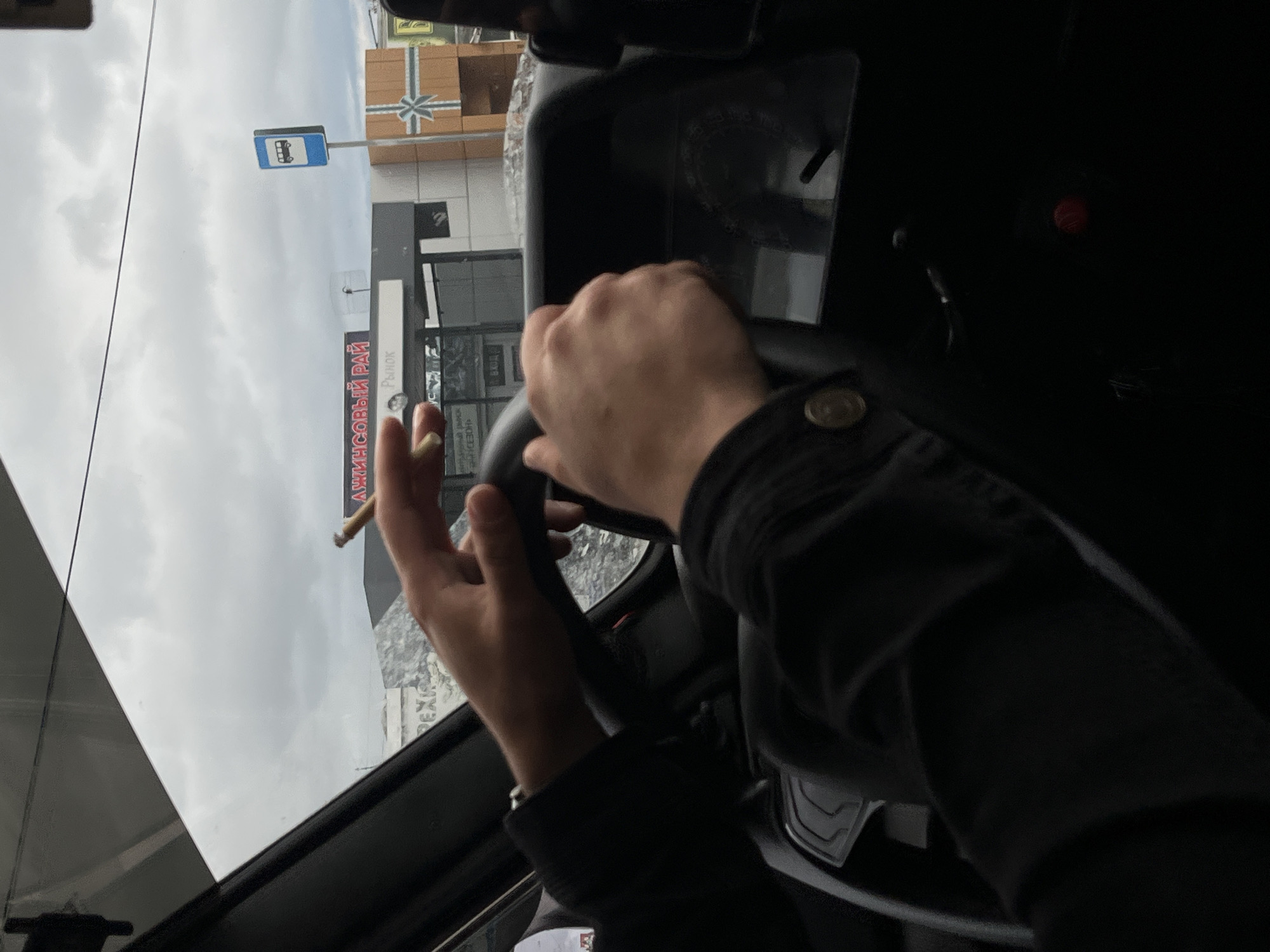 The image shows a person's hands on a steering wheel inside a vehicle, with a cigarette burning between their fingers. The hands are wearing dark clothing. Outside the vehicle, through the windshield, a cloudy sky is visible. In the background, a bus stop sign and a building with signage can be seen. The signage on the building is in Russian and appears to advertise "ДЖИНСОВЫЙ РАЙ" (Dzhinsovyy Ray - Jeans Paradise) and "Рынок" (Rynok - Market). The location is identified as Chaikovsky, Russia.