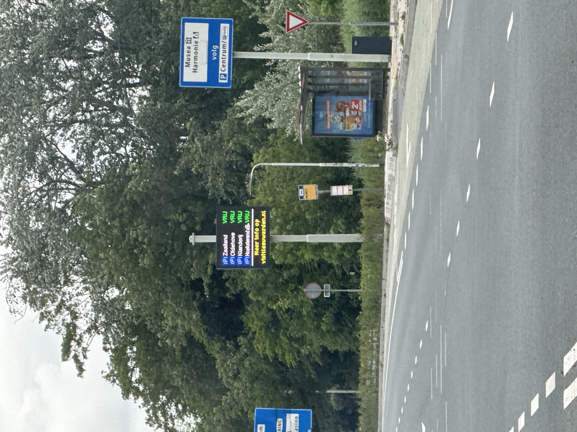 This image depicts a road scene in Leeuwarden, Netherlands, with a focus on traffic and information signage. In the foreground, a two-lane road with white dashed and solid lines is visible. On the right side of the road, a bus stop shelter with advertisements is present, along with a dark blue trash bin and a white pole. Adjacent to the bus stop is a tall pole with a digital display showing parking information for various locations in and around Leeuwarden, such as Zaailand, Oldehove, and Klanderij, indicating "VRIJ" (free). Below this, it directs users to visitleeuwarden.nl for more information. Further right, a white triangular yield sign is positioned. In the background, dense green trees and foliage line the road, with a cloudy sky above. A blue sign with white lettering indicating "Musea Harmonie" and "volg P Centrum/route" is also visible. A traffic light or signal is visible in the middle ground.