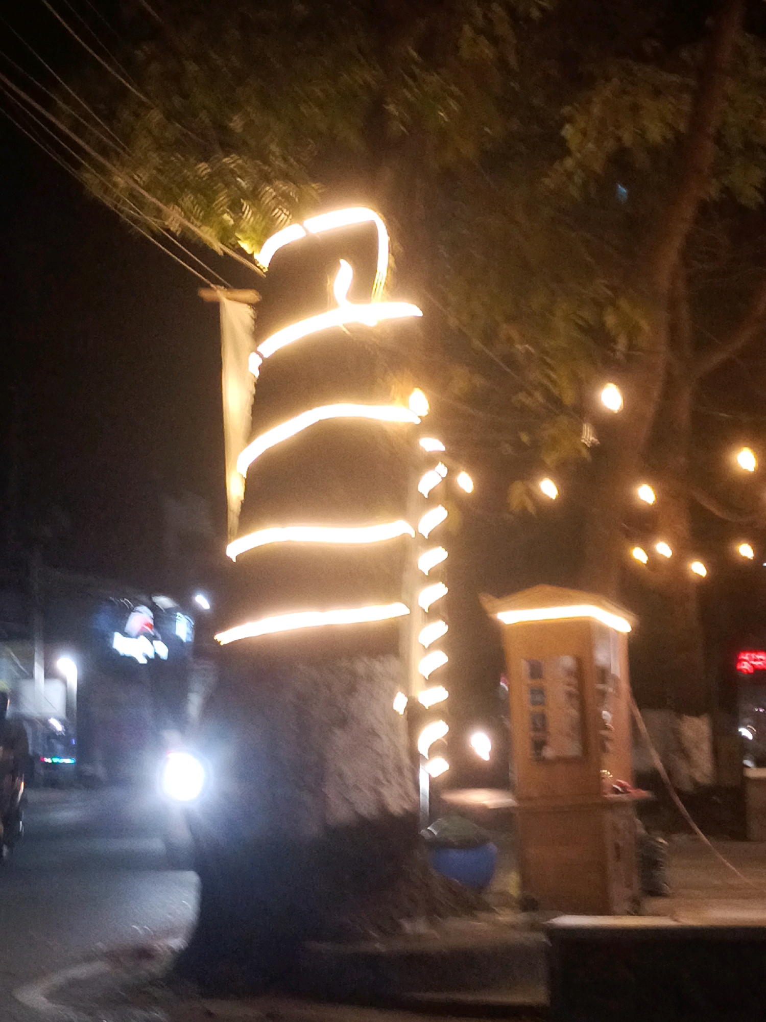 A nighttime street scene in Malang, Indonesia, is depicted, featuring a tree wrapped in bright, festive string lights. These lights create a spiral pattern around the trunk, emitting a warm, yellow glow. In the background, out-of-focus trees with further string lights suggest a public space or event. A small, illuminated wooden kiosk-like structure stands to the right of the tree. A motorcycle with a bright headlight is partially visible in the lower left of the frame, suggesting ongoing traffic. The overall atmosphere is one of a decorated urban environment at night.