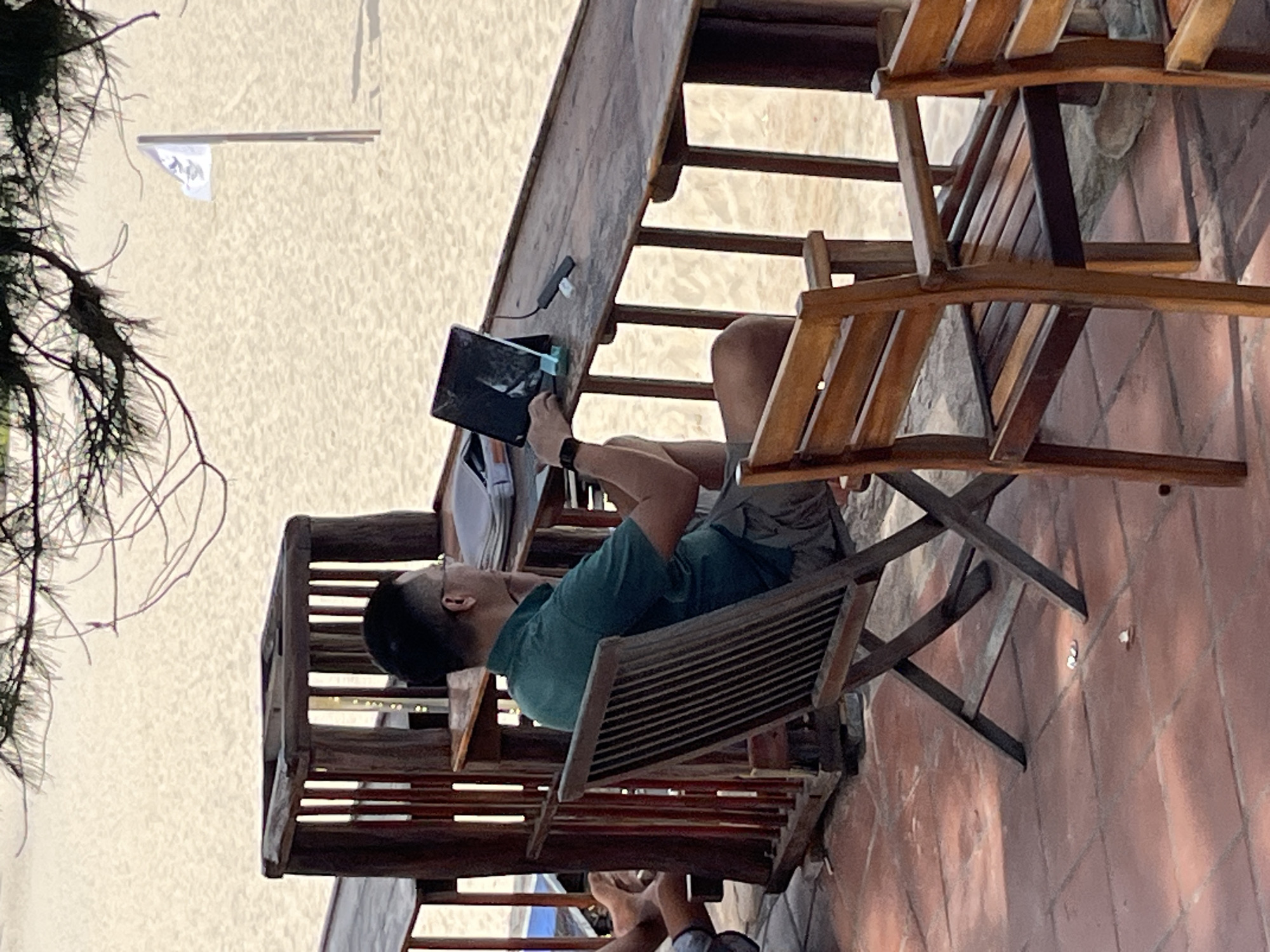 A man in a green polo shirt and khaki shorts is sitting at a wooden table on a tiled patio overlooking a sandy beach. He is wearing glasses and a watch on his left wrist and is looking at a tablet that is propped up on the table. The tablet is plugged into a charging cable. To the left of the man, there is a wooden chair with a person's legs visible. In the background, there is a white flag on a pole planted in the sand, and pine trees branch over the scene. The location is a beach cafe in Mũi Né, Vietnam.