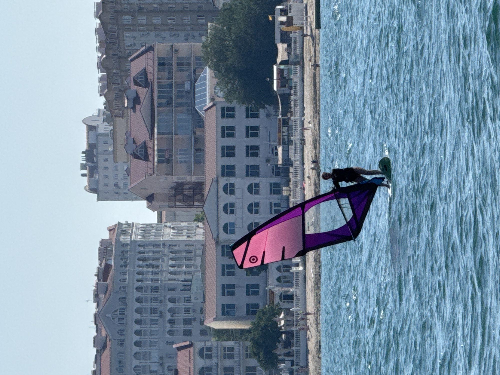 A person is windsurfing on the Black Sea in front of a coastal city. The person is standing on a board with a sail attached, which is colored pink and purple. The water is choppy, and the sky is clear and blue. In the background, there are several large buildings with a distinctive architectural style, including arched windows and ornate facades. These buildings are characteristic of the historical architecture found in Sevastopol, Ukraine. The beachfront is visible with some people walking and sitting on the sand. The scene captures a leisure activity against the backdrop of an urban coastal landscape.