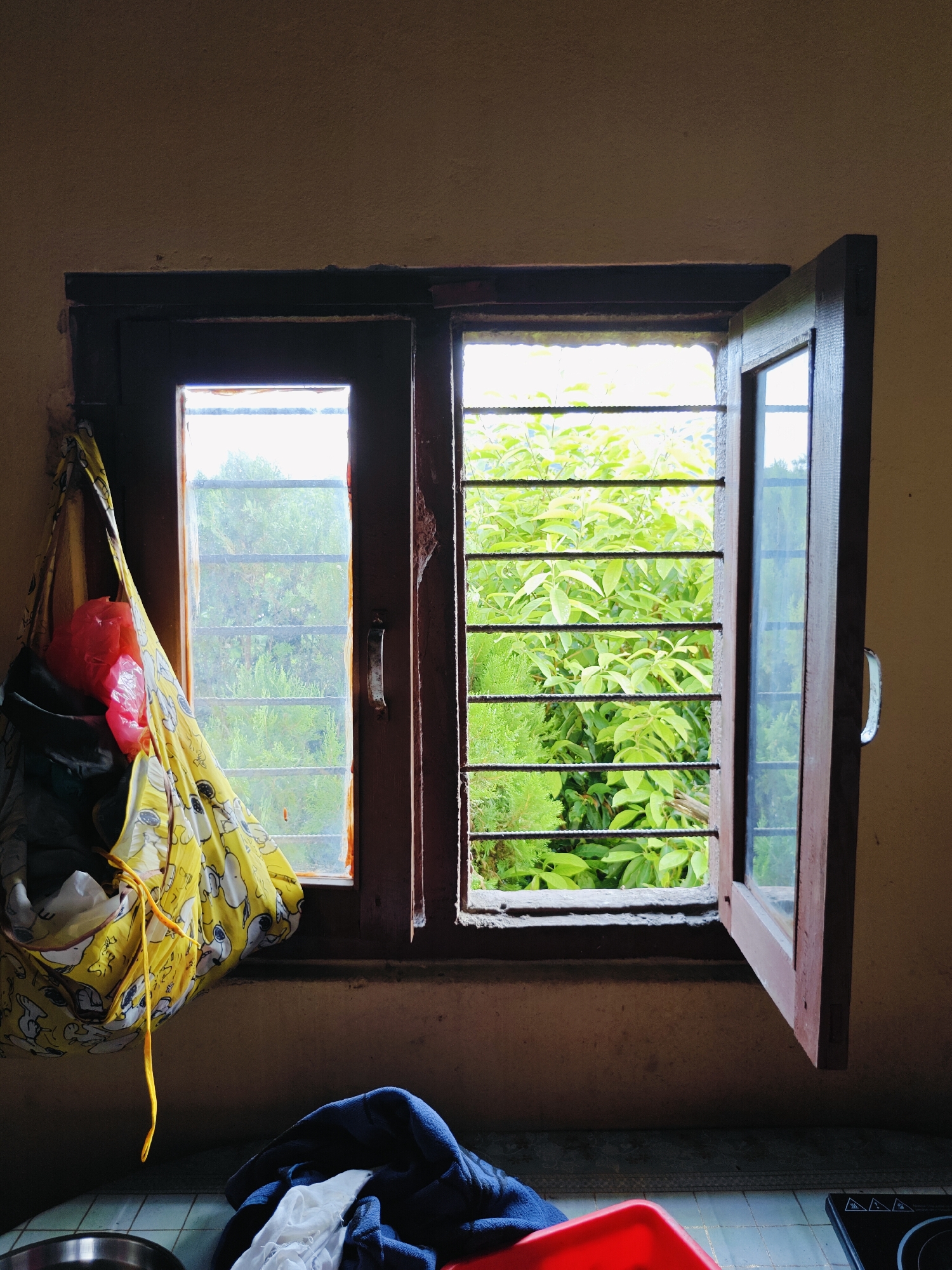 The image depicts an interior scene looking out a window in Pokhara, Nepal. The window has a wooden frame and is divided into two panes. The left pane is mostly covered by a yellow fabric bag with a cartoon print, which is hanging from something outside the frame and contains a red plastic bag and other items. The right pane is open, revealing a lush green outdoor environment with dense foliage and trees. Horizontal bars are visible across the right pane. Inside, on the windowsill and counter below, are some crumpled pieces of fabric, including a blue and white garment, and a red plastic container. The walls appear to be textured and light brown. The overall impression is of a simple, possibly domestic, setting with a view of abundant nature outside.