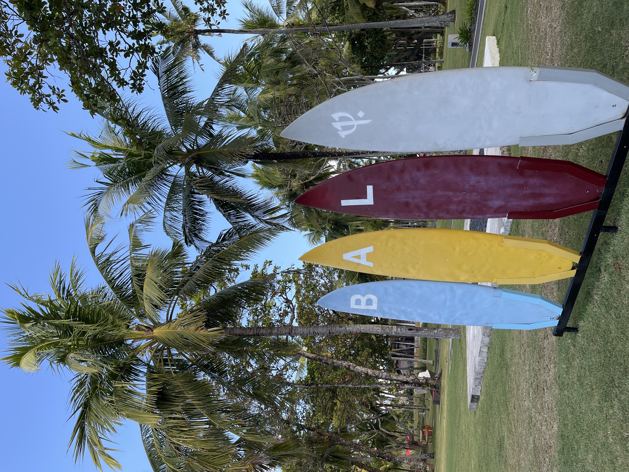 In Kuta Selatan, Indonesia, four surfboards stand upright on a grassy area beneath palm trees. The surfboards are colored light blue, yellow, maroon, and gray, with letters and symbols painted on them. The light blue board has a white "B", the yellow board has a white "A", and the maroon board has a white "L". The gray surfboard has a white trident symbol. In the background, there are more palm trees, other foliage, and some structures that appear to be buildings, possibly part of a resort or restaurant, with a clear blue sky above. The surfboards are arranged in a row and are attached to a black metal stand. The area appears to be well-maintained, with neatly cut grass. There are no people visible in the immediate foreground or interacting with the surfboards.