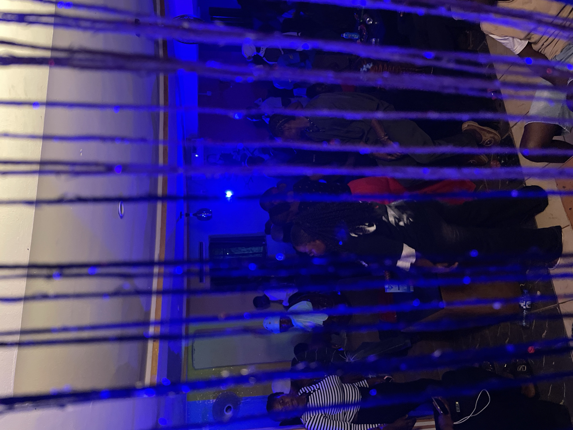 A dimly lit indoor scene, likely a party or gathering, is captured through a curtain of blue, hanging strands. The environment appears to be a room with white walls and a ceiling adorned with what seem to be decorative lights or bulbs, some of which emit a blue glow. In the background, several individuals are visible, engaged in various activities. On the left, a woman in a black and white striped shirt is looking forward, while another woman with dark curly hair is visible on the right. Other people are scattered throughout the space, some standing and others seated. The overall lighting is low, with prominent blue hues casting a somewhat subdued atmosphere. The specific location in Durimi, Nigeria, is not discernible from the visual information provided.