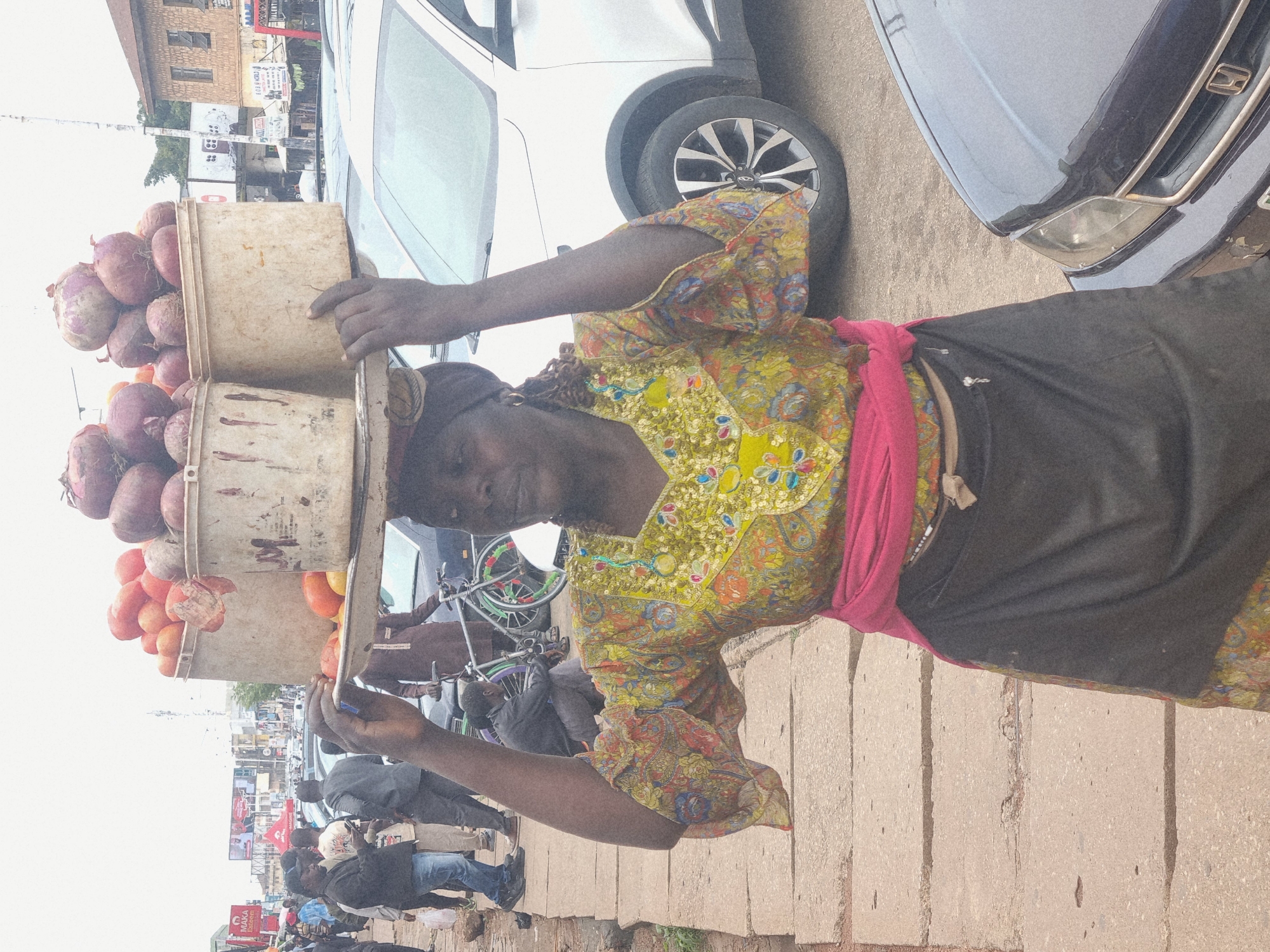 This image captures a street scene in Jos, Nigeria, with a focus on a woman carrying a large load of produce on her head. The woman, dressed in a vibrant, patterned yellow top and a pink scarf, appears to be a vendor. The produce consists of numerous red onions in two stacked containers, likely baskets or buckets, also carrying some smaller, round fruits which appear to be tomatoes or similar. She is walking on a paved sidewalk, with a busy street scene unfolding behind her.
In the background, a variety of vehicles, including cars and possibly a motorcycle, are visible, suggesting a moderately trafficked area. The architecture of the buildings in the background, with their brickwork and windows, hints at a commercial or residential district. The sky is overcast, suggesting it might be a cloudy day or late afternoon. The overall impression is of a typical day in a bustling Nigerian city, where street vending is a common practice.
Notable details include the woman's determined posture as she balances the heavy load, a testament to her livelihood. The detail of her colorful attire contrasts with the more subdued colors of the surroundings. The texture of the paved sidewalk and the various vehicles provide a sense of the urban environment. No discernible text is clearly legible in the image.