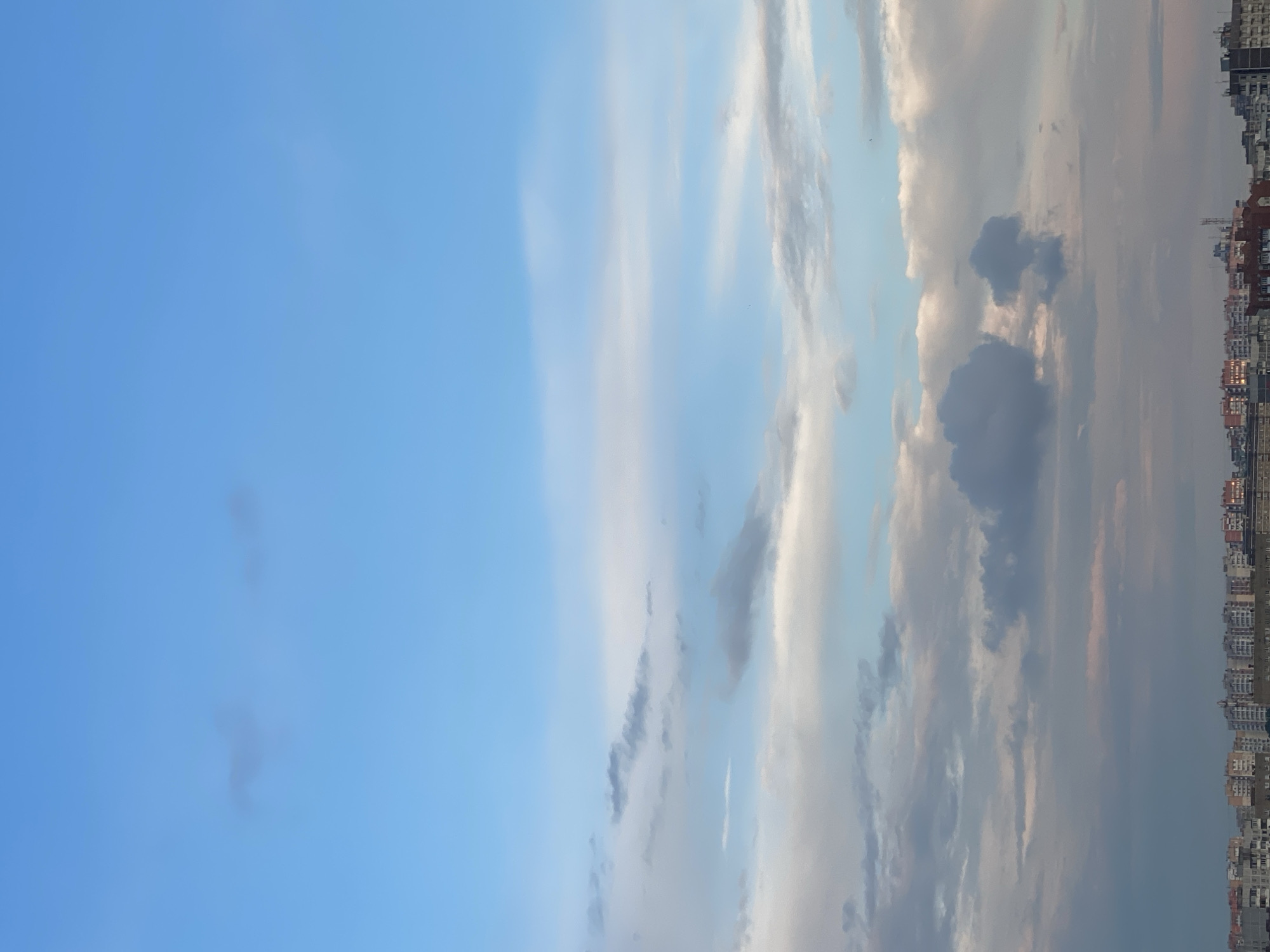 The image displays a cityscape beneath a vast, pale blue sky filled with scattered clouds of varying density and color. The clouds range from wispy, thin streaks of white to darker, more voluminous grey masses. In the distance, the horizon is lined with a dense collection of low-rise buildings, characterized by multiple stories, balconies, and a variety of muted colors, including shades of grey, white, orange, and red. The architecture appears to be residential or commercial, with uniform rooflines and window patterns suggesting a developed urban area. The lighting suggests it might be late afternoon or early evening.