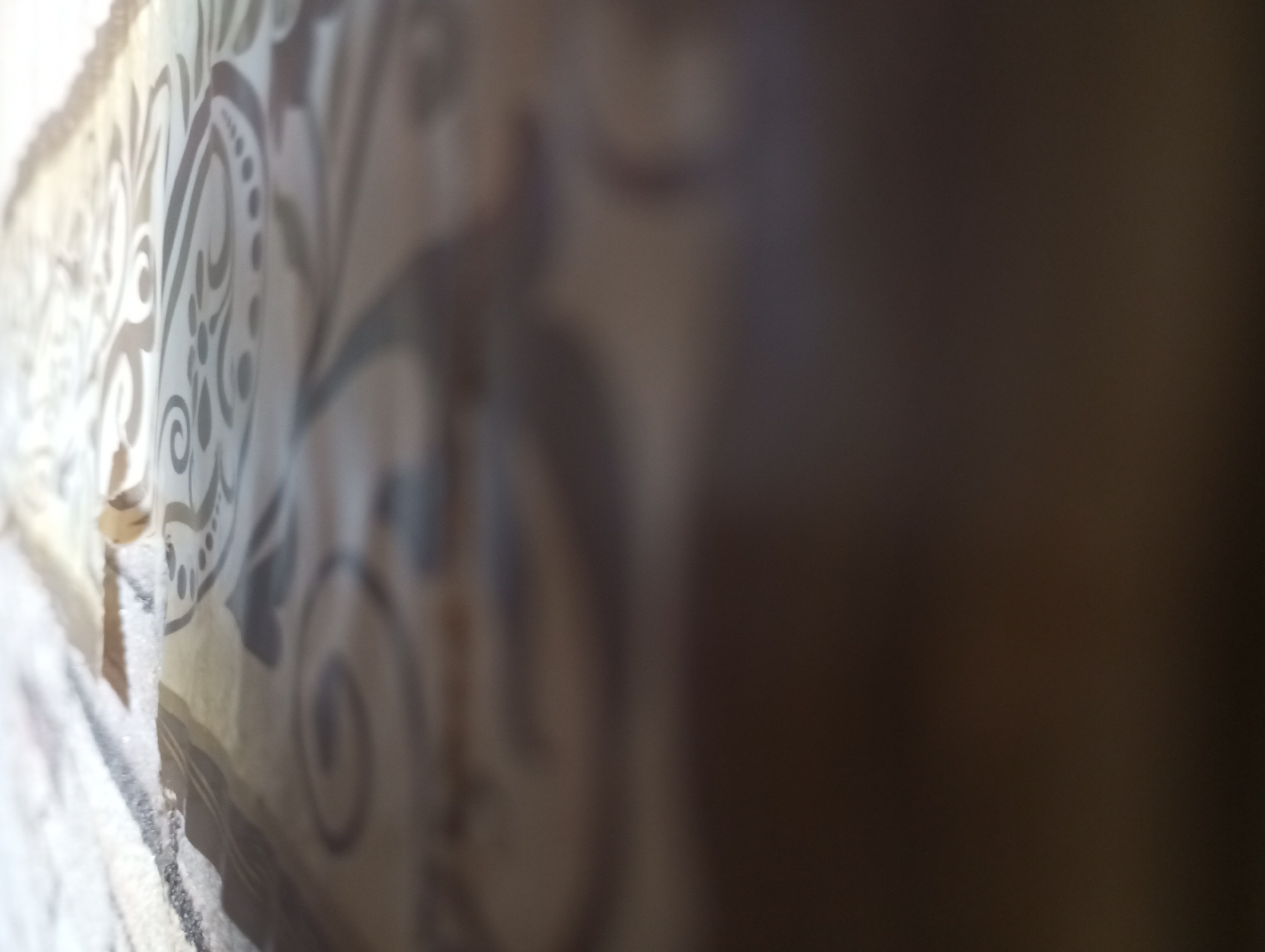The image depicts a close-up, out-of-focus view of a patterned fabric. The pattern appears to be a repeating, ornate design with swirling lines and dots, possibly a damask or brocade style. The colors are muted, with shades of beige, brown, and off-white. The lighting suggests it is indoors, with a light source coming from the left, casting a slight glare on the fabric's surface. The right side of the image is darker and significantly blurred, indicating a separation or a different material further away from the camera. There is no discernible information about the city of Kirzhach, Russia, or any people or specific actions within this image. The scene is an abstract close-up of textile texture and pattern.