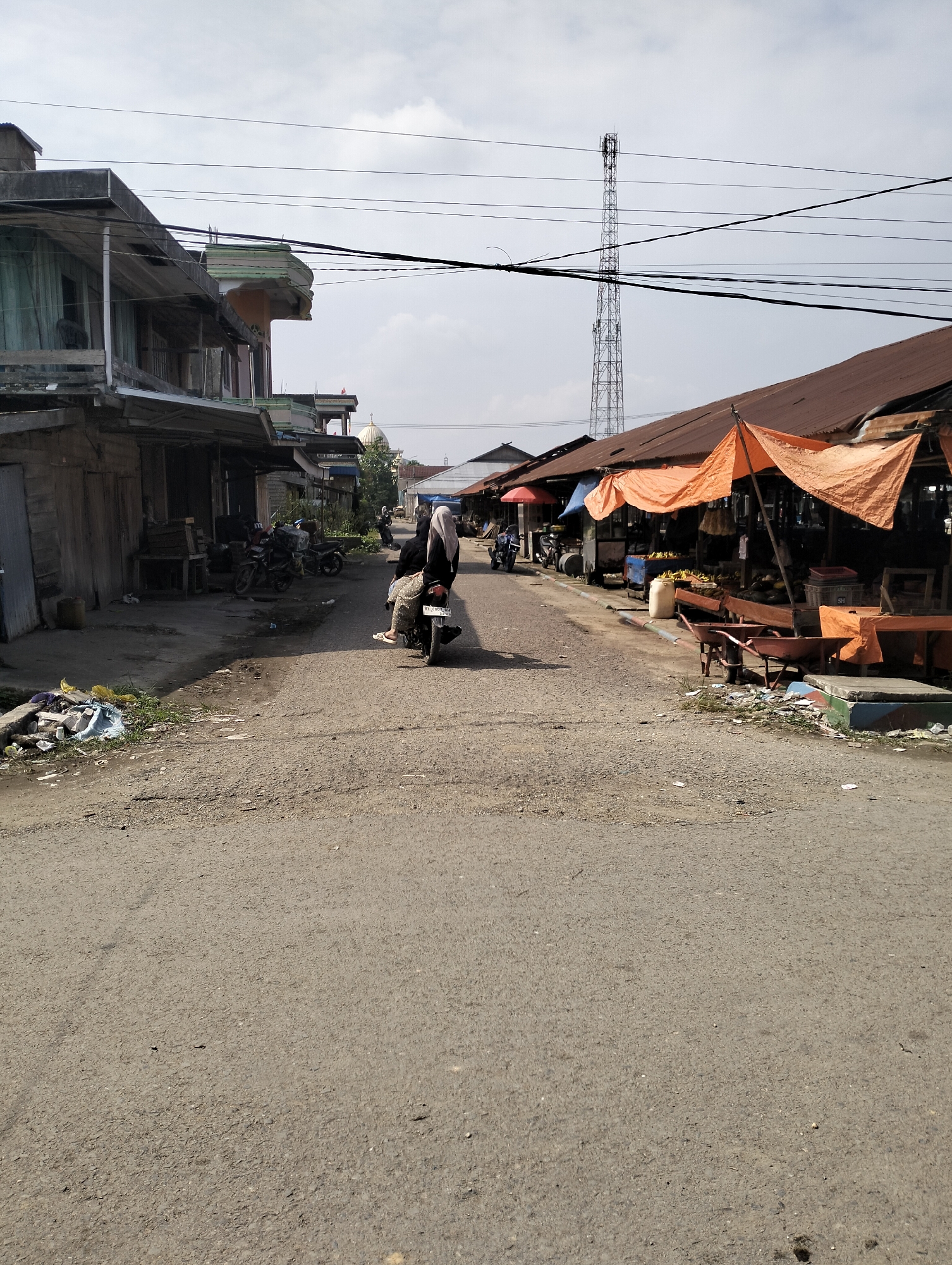 The image depicts a street scene in Serdang Jaya, Indonesia, likely a local market area. A person on a motorcycle, wearing a headscarf and patterned skirt, is the central moving subject, traveling down the asphalt road. On the left, a row of aged buildings with wooden facades and exposed construction elements lines the street, some with covered verandas. On the right, market stalls are set up under orange and blue tarps, displaying produce like bananas and possibly other fruits or vegetables. Several wheelbarrows are visible near the stalls. In the background, a tall metal tower stands against a cloudy sky, and a domed building, likely a mosque, is visible further down the street. Various motorcycles are parked along the left side of the road, and debris is scattered on the ground near the buildings and the road's edge.