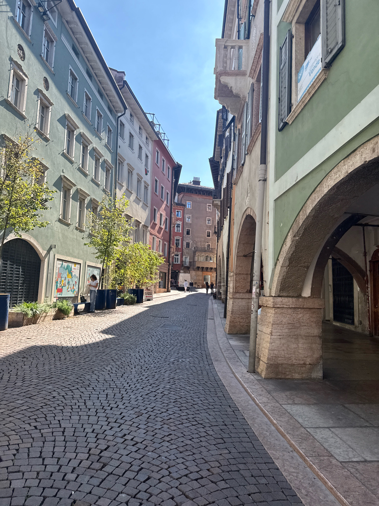 The image depicts a street scene in the city of Trento, Italy. The street is paved with cobblestones and curves to the left. On the left side of the street, a row of colorful buildings with multiple stories and windows is visible. The buildings are painted in shades of green, white, and pink. There are trees planted in large blue containers along the sidewalk in front of the green building. A person is standing near the trees, looking at a large poster. On the right side of the street, there is a covered walkway with arched openings supported by stone pillars. A sign is visible in one of the upper windows of a building on the right, which appears to be a real estate listing. The sky is clear and blue. The sun is casting shadows on the street, indicating it is daytime. Several people are walking in the distance on the street.