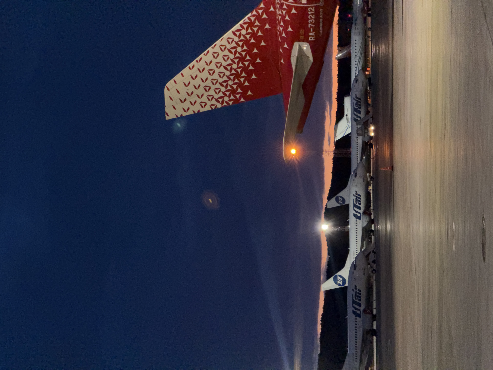 The image shows a row of UTair airplanes parked on an airport tarmac at dusk, with a red and white tail fin of a separate airplane in the foreground. The location is most likely Емельяново аэропорт, Russia. The sky is dark blue, transitioning to orange near the horizon where the sun has set. The airport tarmac is illuminated by overhead lights, reflecting off the wet surface. The UTair airplanes are white with blue lettering. The red and white tail fin has a geometric pattern.