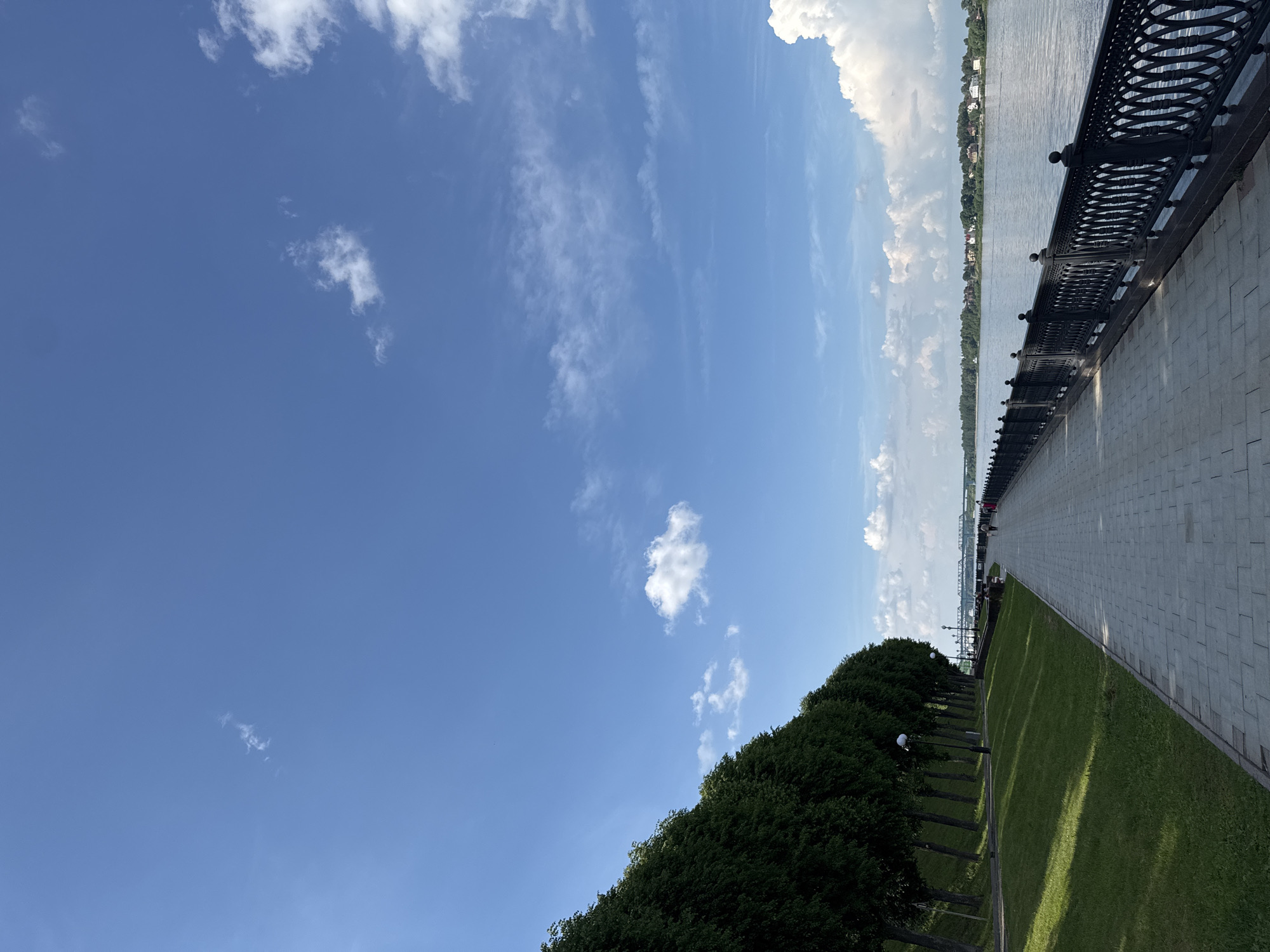 The image depicts a scenic view of the Volga River embankment in Zavolzhye, Russia. A paved walkway lines the river, bordered by an ornate black metal railing. Beyond the railing, the wide, calm river stretches towards a distant treelined shore dotted with houses and buildings. The sky above is a bright blue, with scattered white clouds. On the left side of the walkway, a grassy slope is covered with evenly spaced, lush green trees. A few people are visible walking along the embankment in the distance. The scene is bathed in sunlight, casting shadows across the walkway and the grassy slope, suggesting a pleasant day.